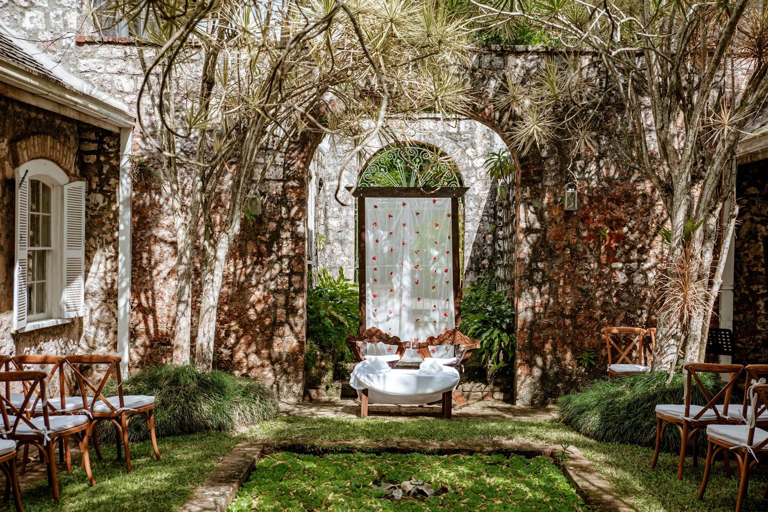 Rustic stone courtyard with arched doorways, lush climbing vines, wooden chairs, and an elegant white table centerpiece.
