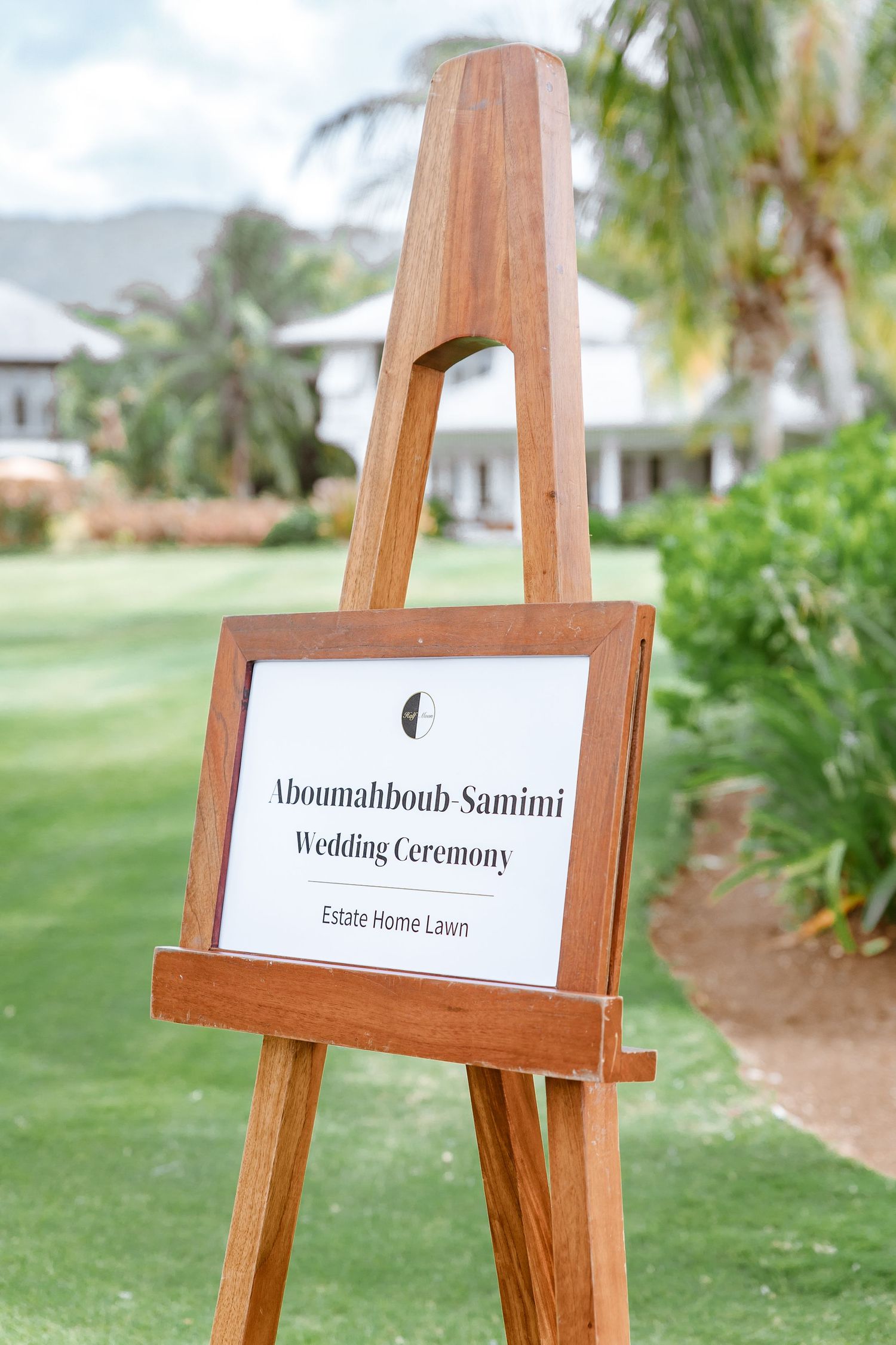 Wooden easel sign for Aboumahboub Samimi Wedding Ceremony at Estate Home Lawn surrounded by lush greenery.