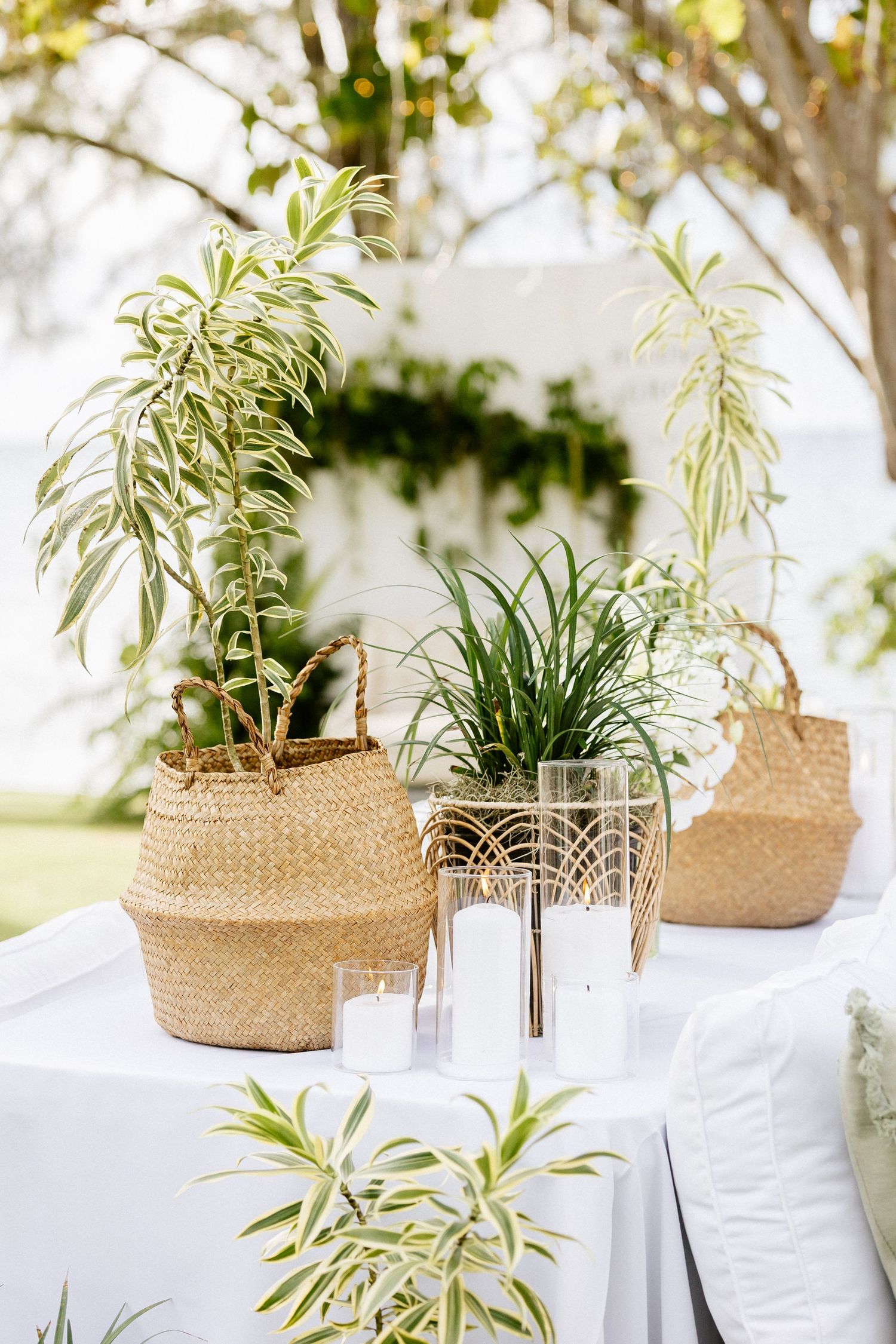 Wicker basket planters with tropical greenery and white candles on a draped table at an outdoor wedding.