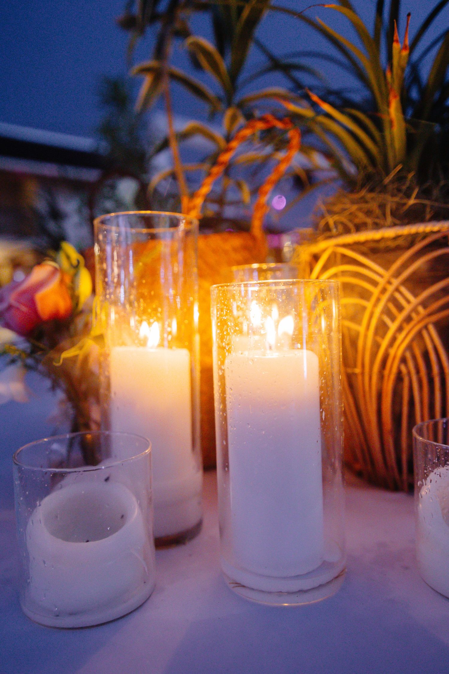 Glowing white pillar candles in glass holders on a table with tropical plants at an outdoor evening event.