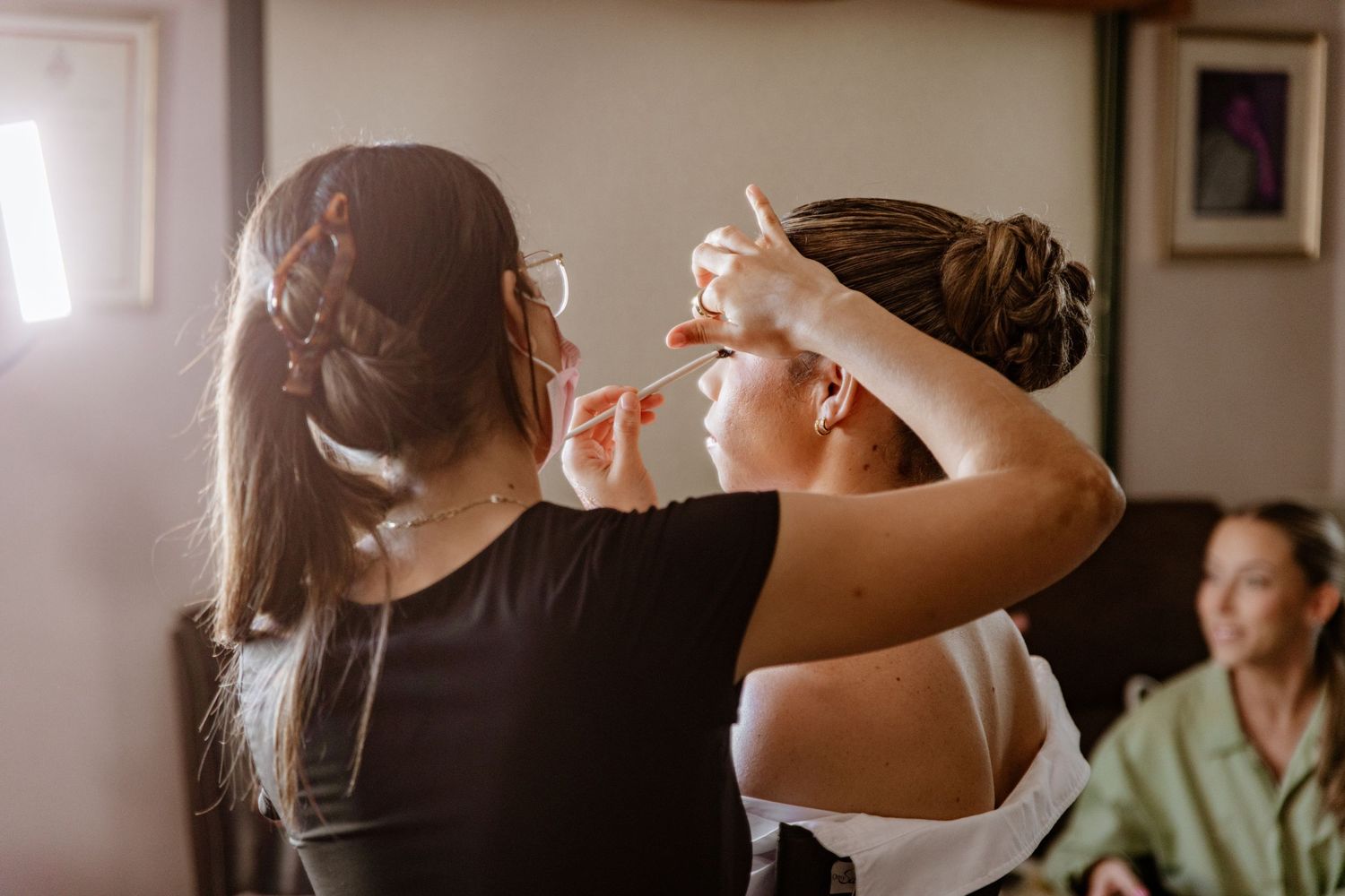 Makeup artist applying eye makeup to bride in bright natural light before wedding.