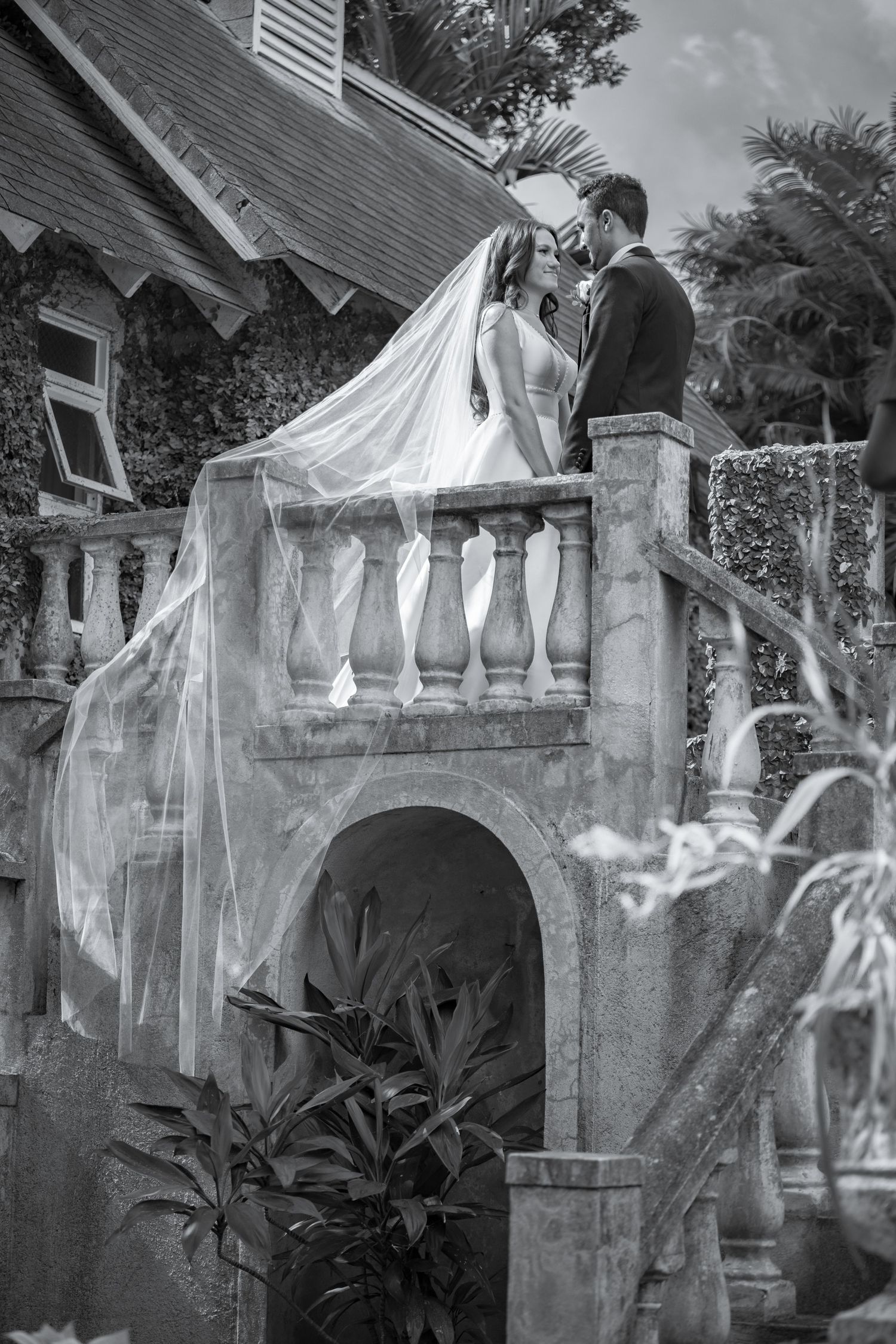 Black and white photo of bride and groom kissing on elegant stone balcony at wedding venue.