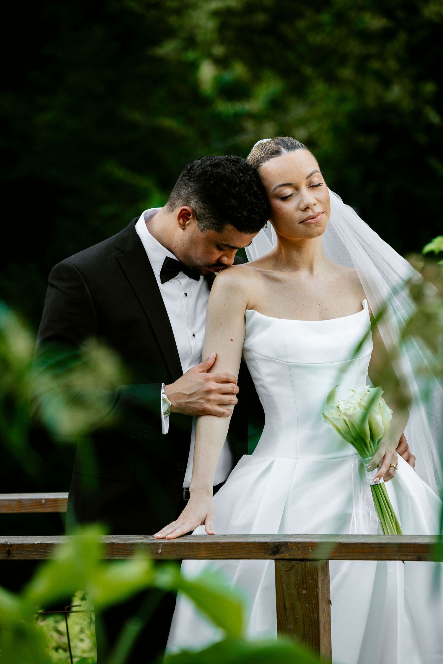 Groom tenderly kisses bride's arm as she leans back gracefully on a wooden bridge in a garden setting.
