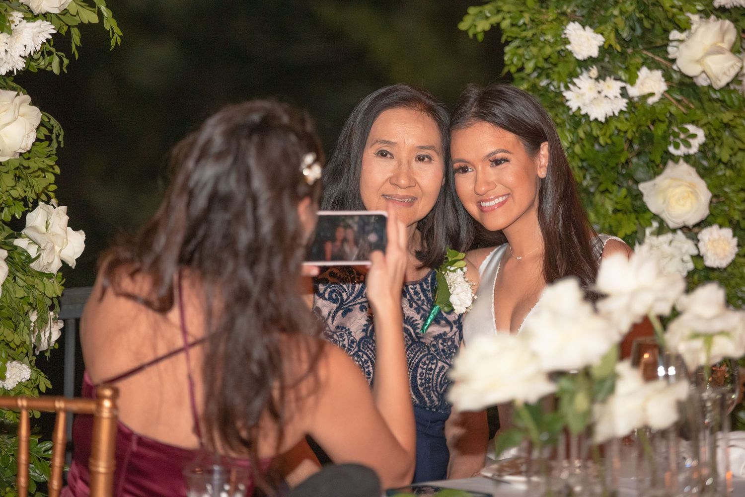 Wedding guests posing for a photo under a lush white floral arch at an outdoor reception dinner.