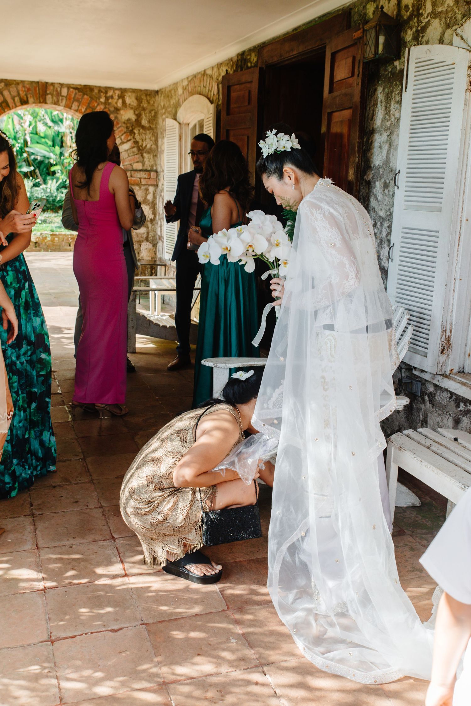 Wedding helper adjusting brides white lace gown while guests in colorful dresses gather at stone venue.