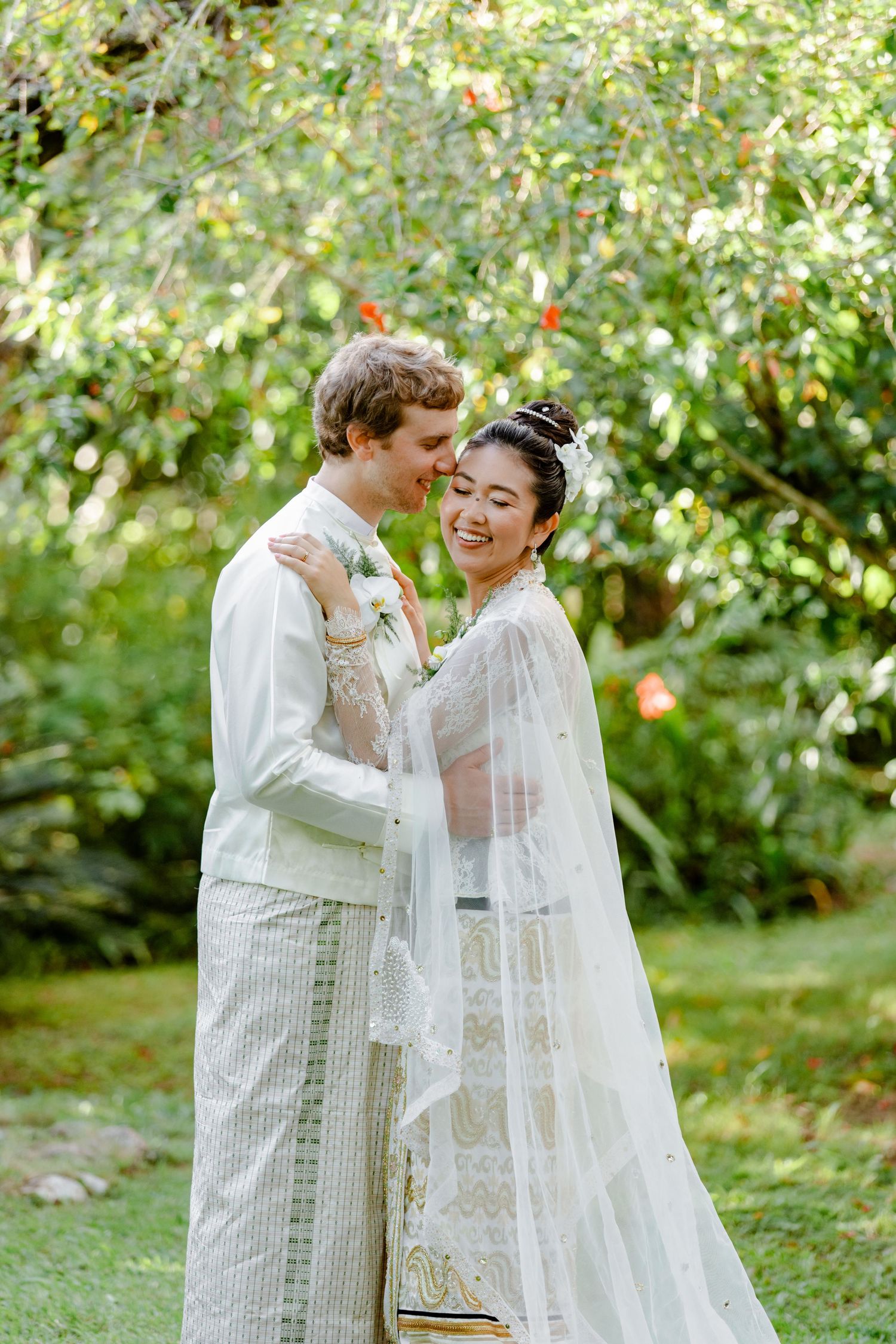 Interracial bride and groom embracing in lush garden wearing traditional Thai wedding attire.