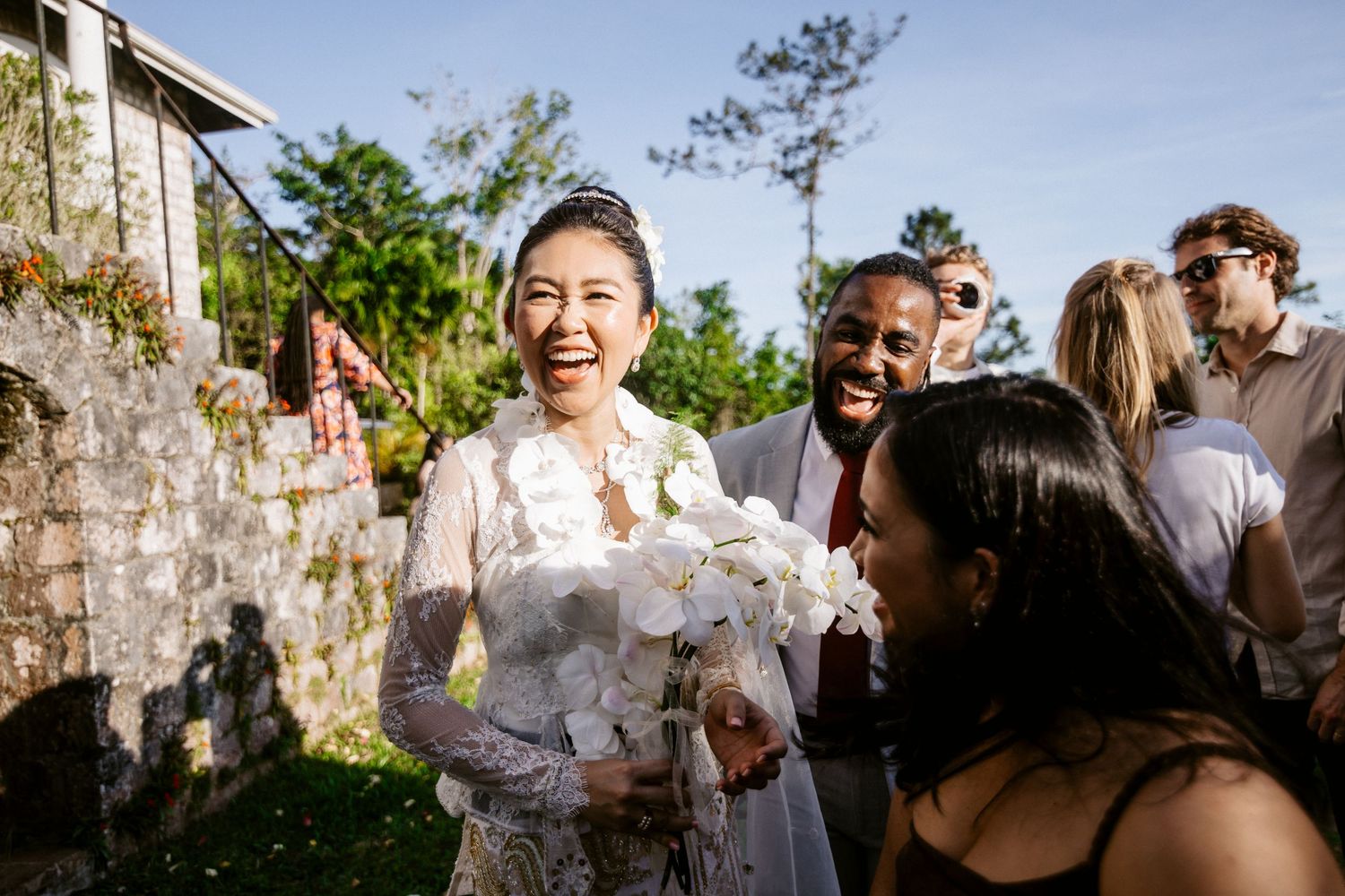 Joyful Asian bride in white lace gown laughing with wedding guests outdoors holding white orchid bouquet.