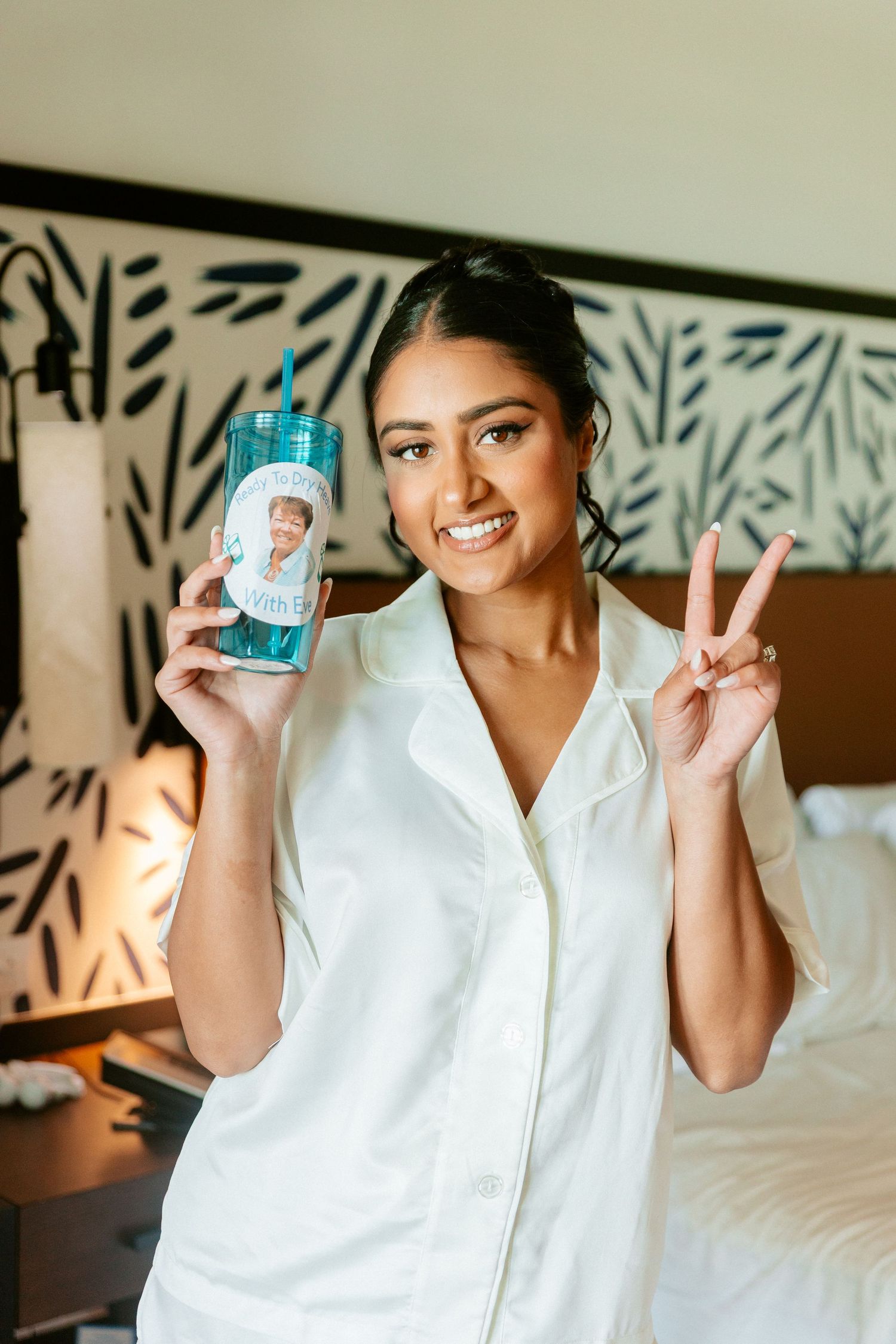 Smiling bride in white pajamas holds a custom tumbler and makes a peace sign during wedding morning preparations.