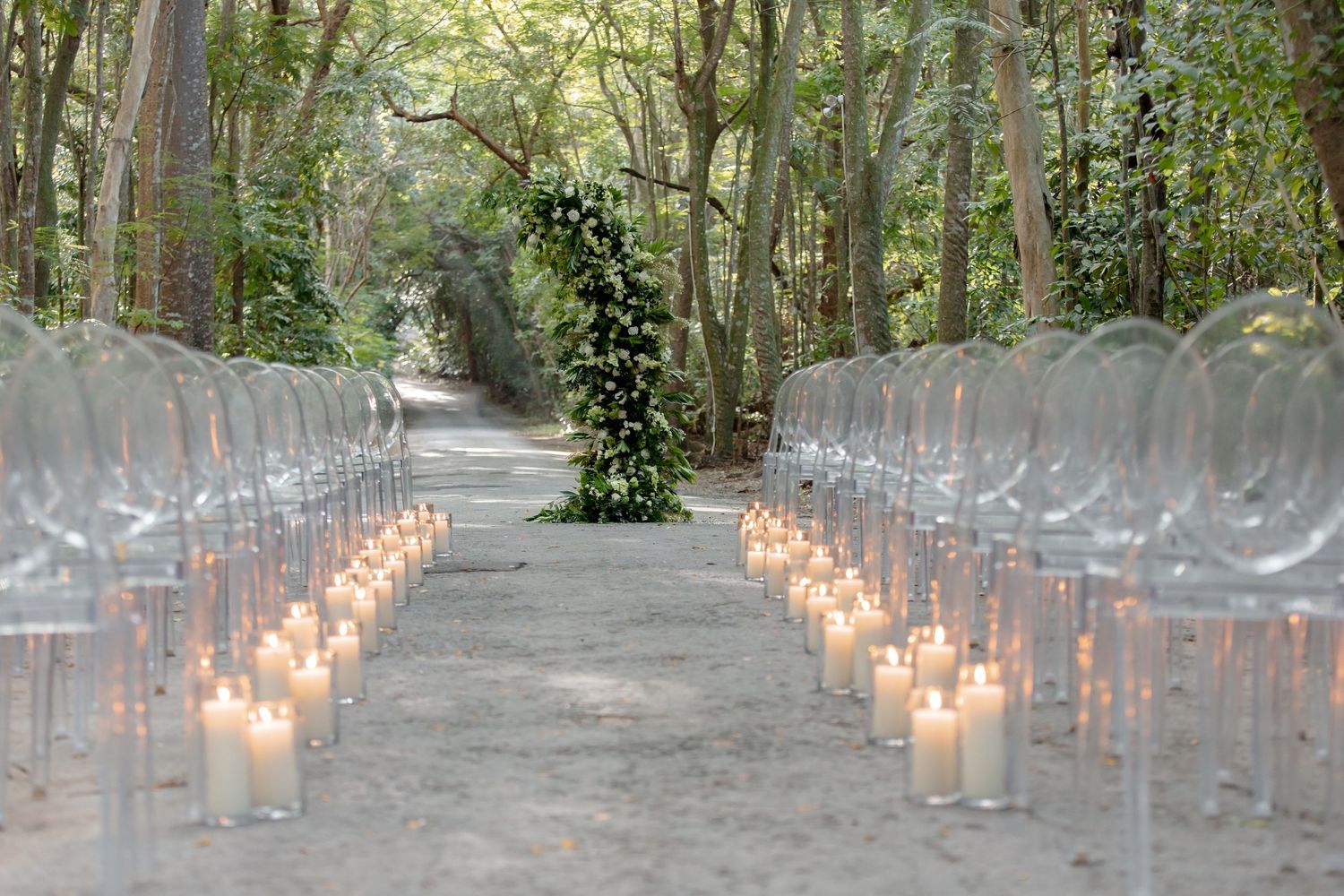 Elegant outdoor wedding aisle with ghost chairs and glowing pillar candles lining a sandy path through a lush green forest.