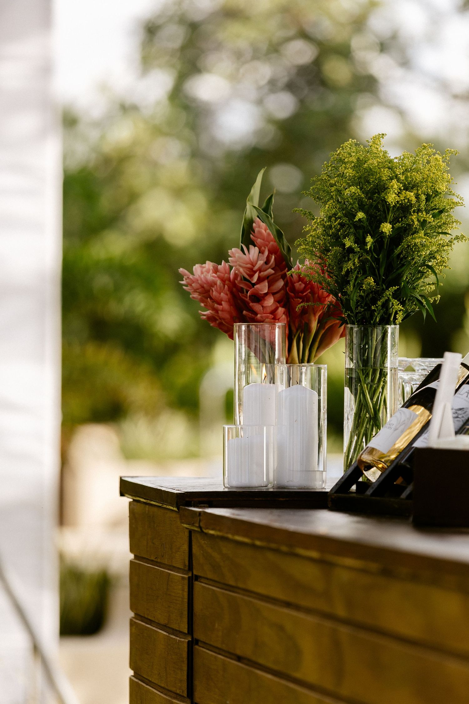 Glass vases with pink ginger flowers and green foliage arranged on a dark wooden cabinet outdoors.