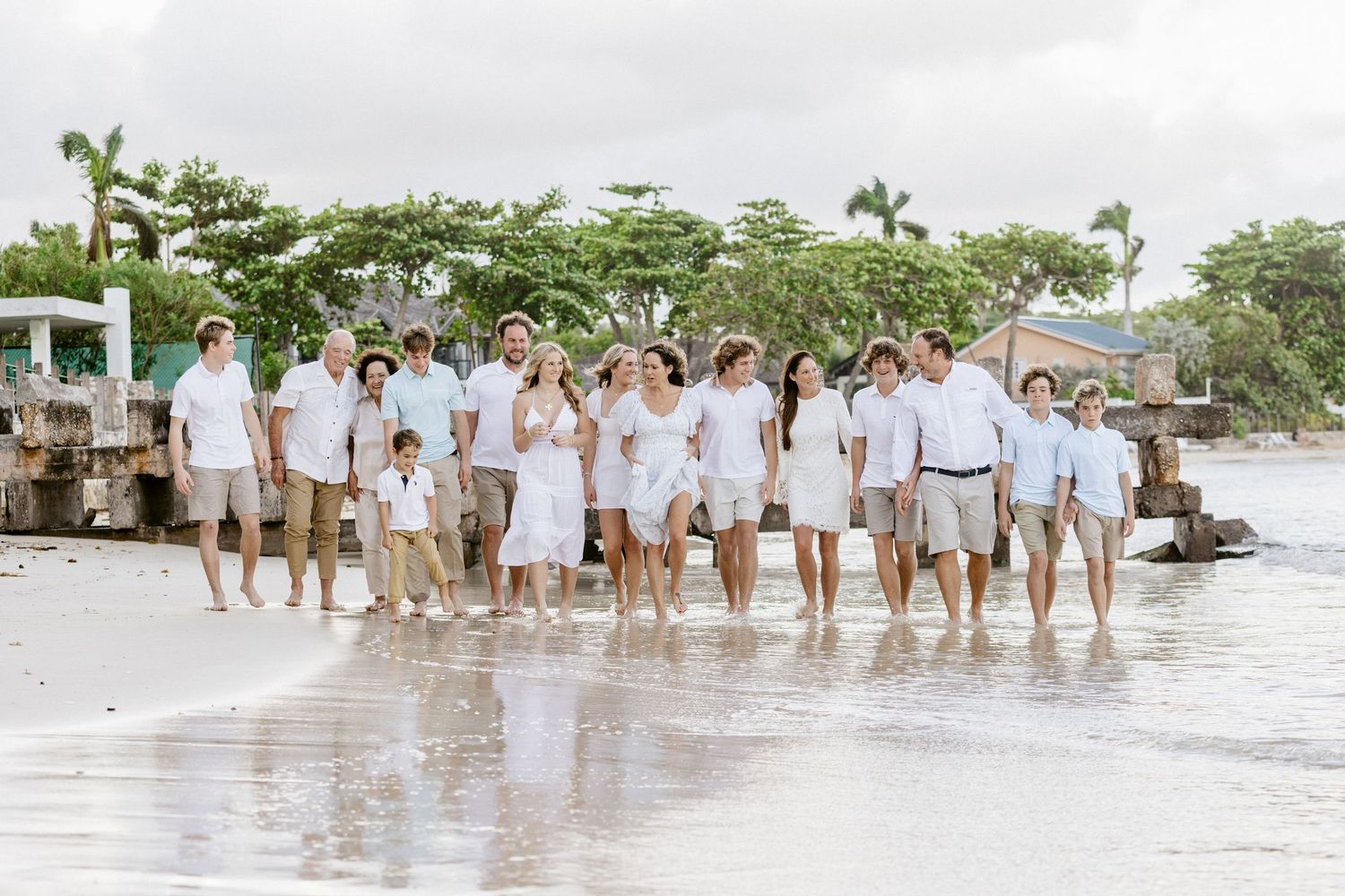Large family group in white and beige outfits posing together on a tropical beach shore with lush green trees behind.