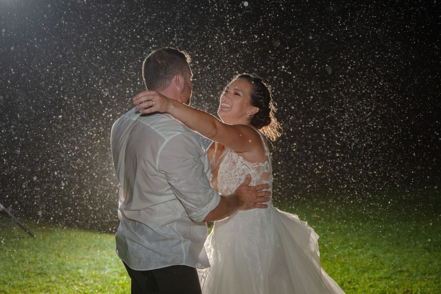 Bride and groom dancing joyfully together in the rain at night during their outdoor wedding celebration.