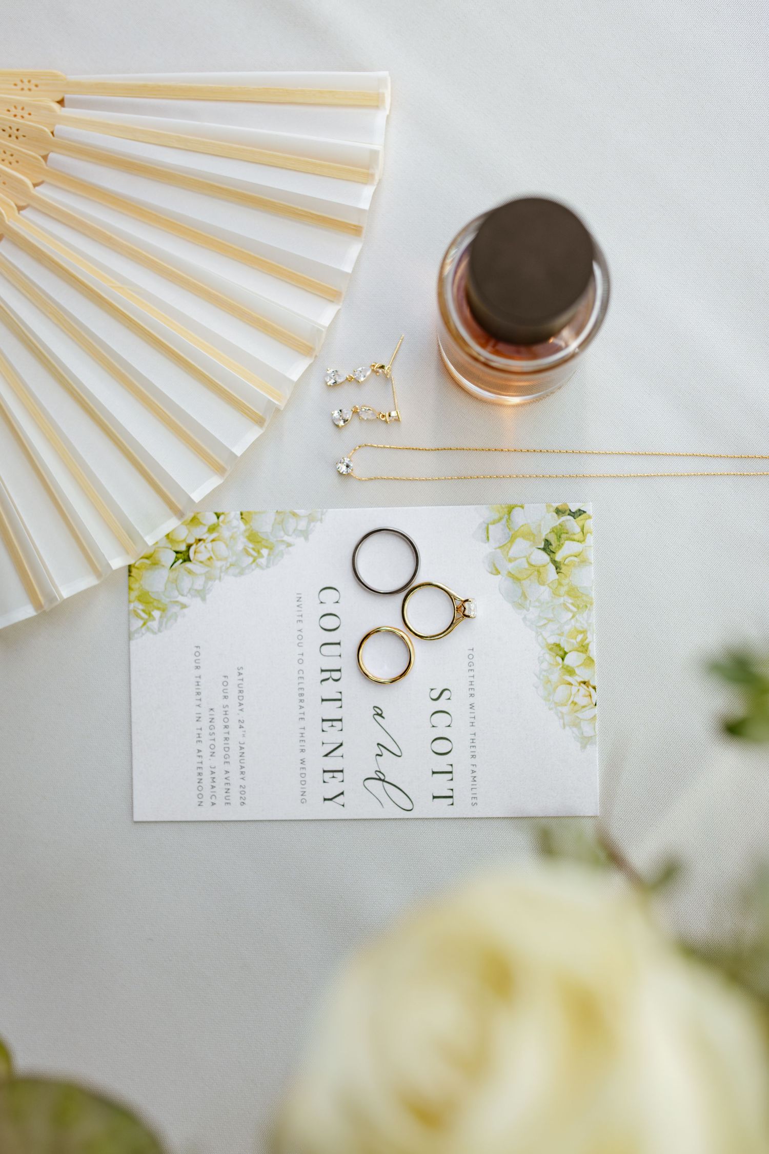 Wedding flat lay featuring gold rings, floral invitation for Scott and Courteney, paper fan, and perfume bottle.