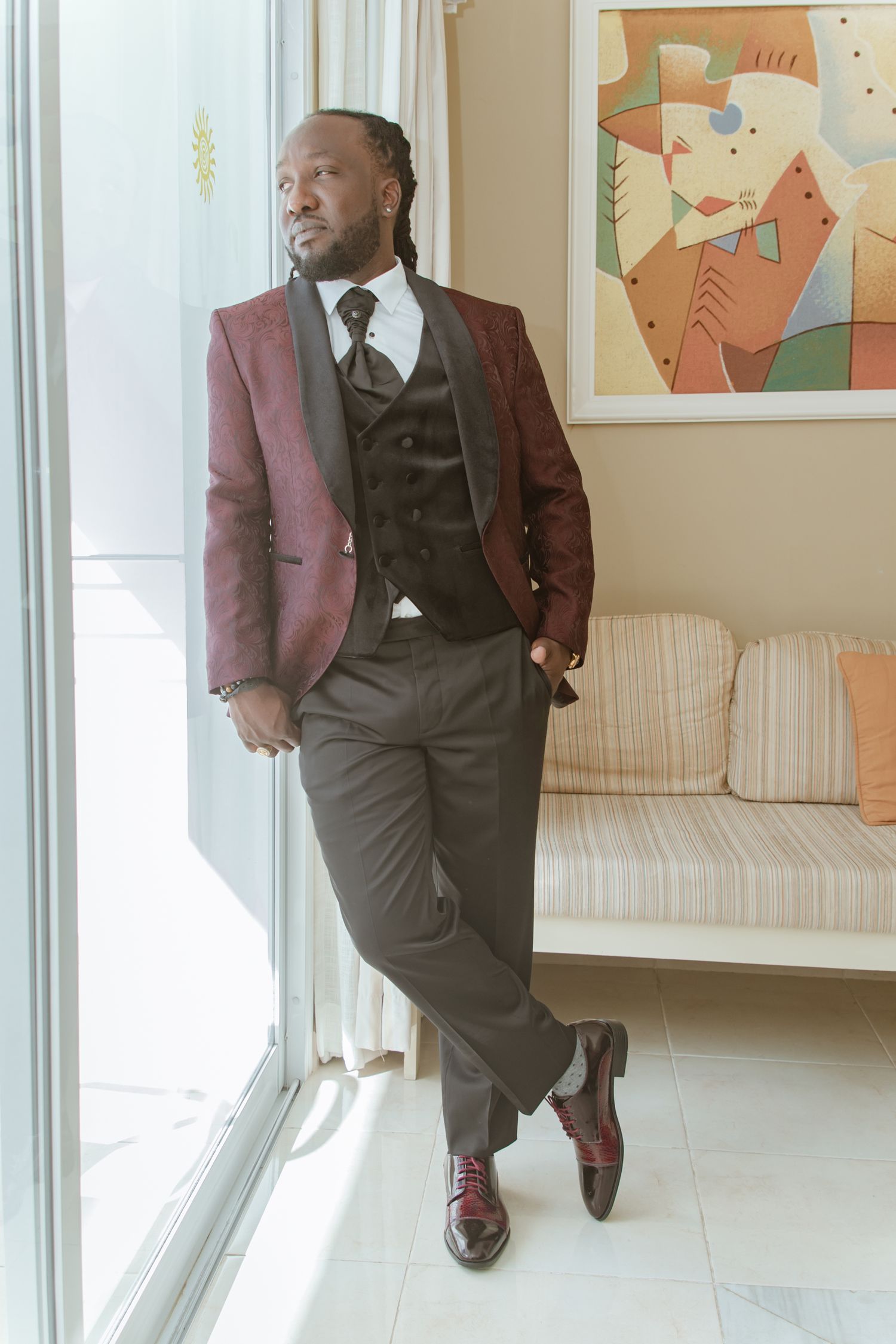 Groom posing confidently in burgundy three-piece suit with dark vest and burgundy dress shoes.