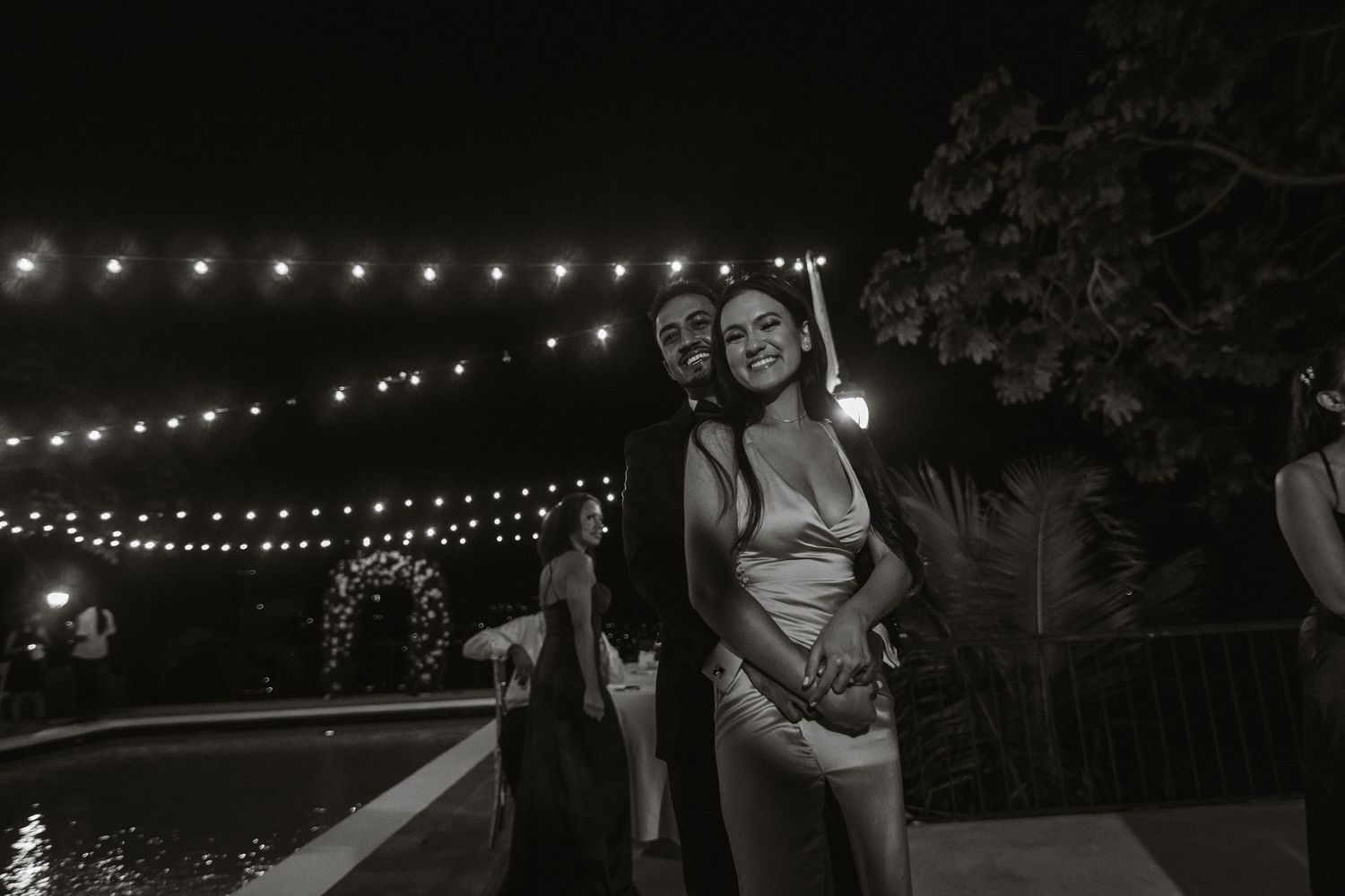 Black and white photo of couple embracing and smiling poolside under string lights at wedding reception.