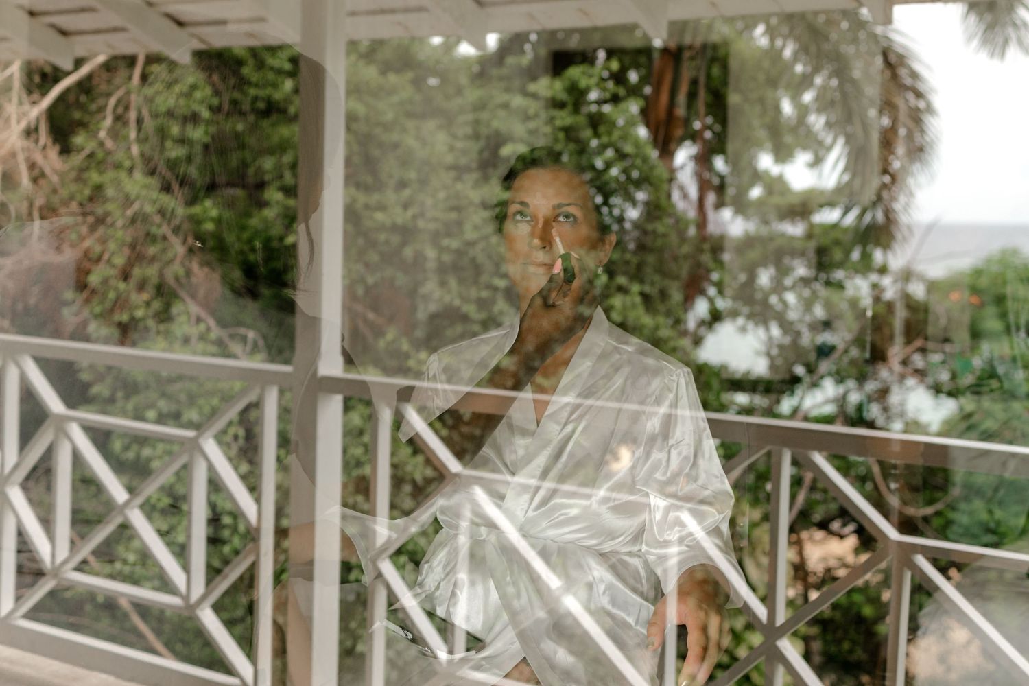 Bride in white robe gazing through glass balcony doors surrounded by greenery.