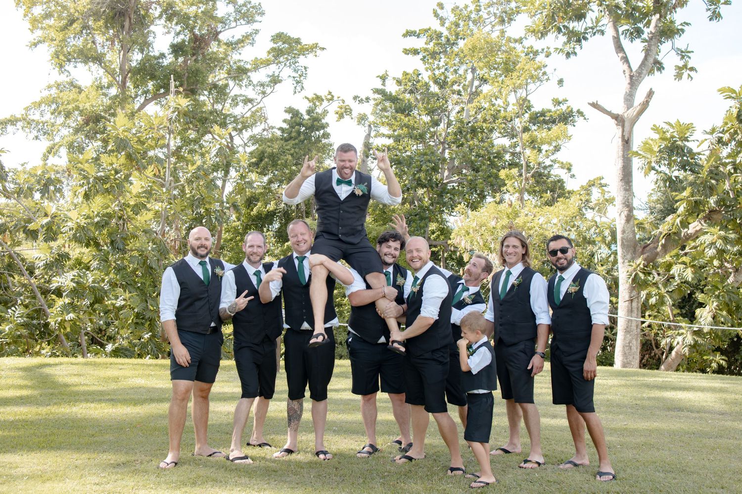 Groomsmen in black vests lifting groom celebrating outdoors surrounded by tropical greenery.