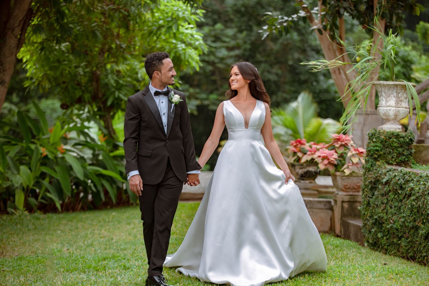 Happy bride and groom walk hand in hand through a green garden with pink flowers.