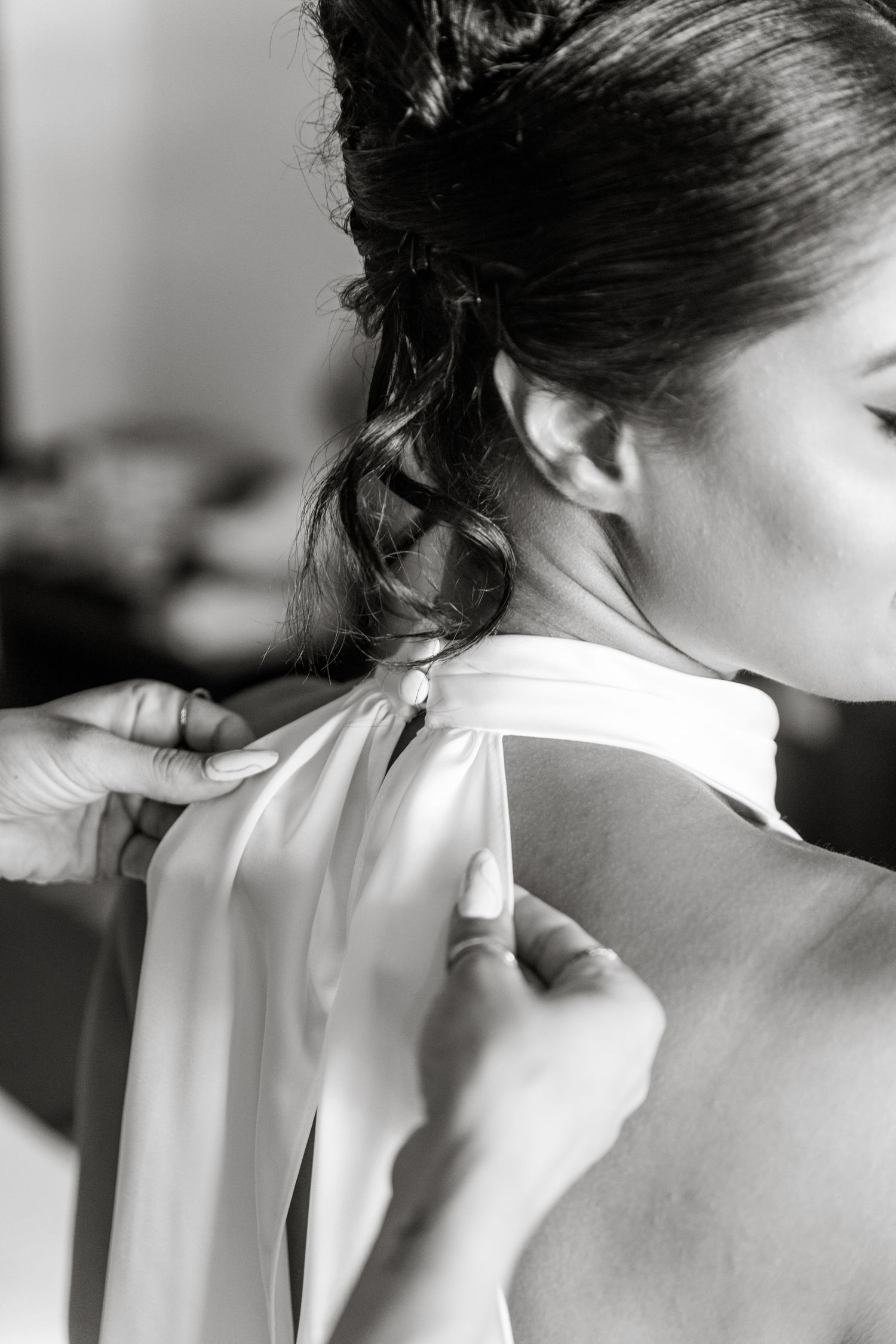 Black and white photo of a bride having her halter wedding dress fastened at the neck by a helper.