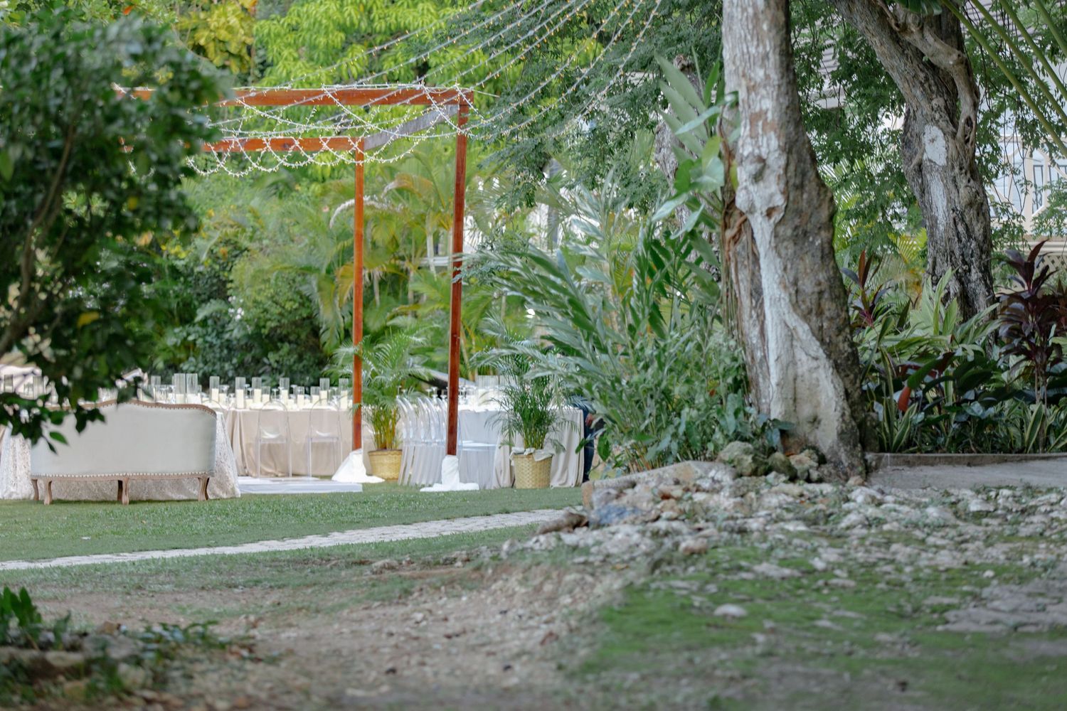 Lush tropical garden with orange metal arch structure, large trees, and green lawn in a serene outdoor setting.