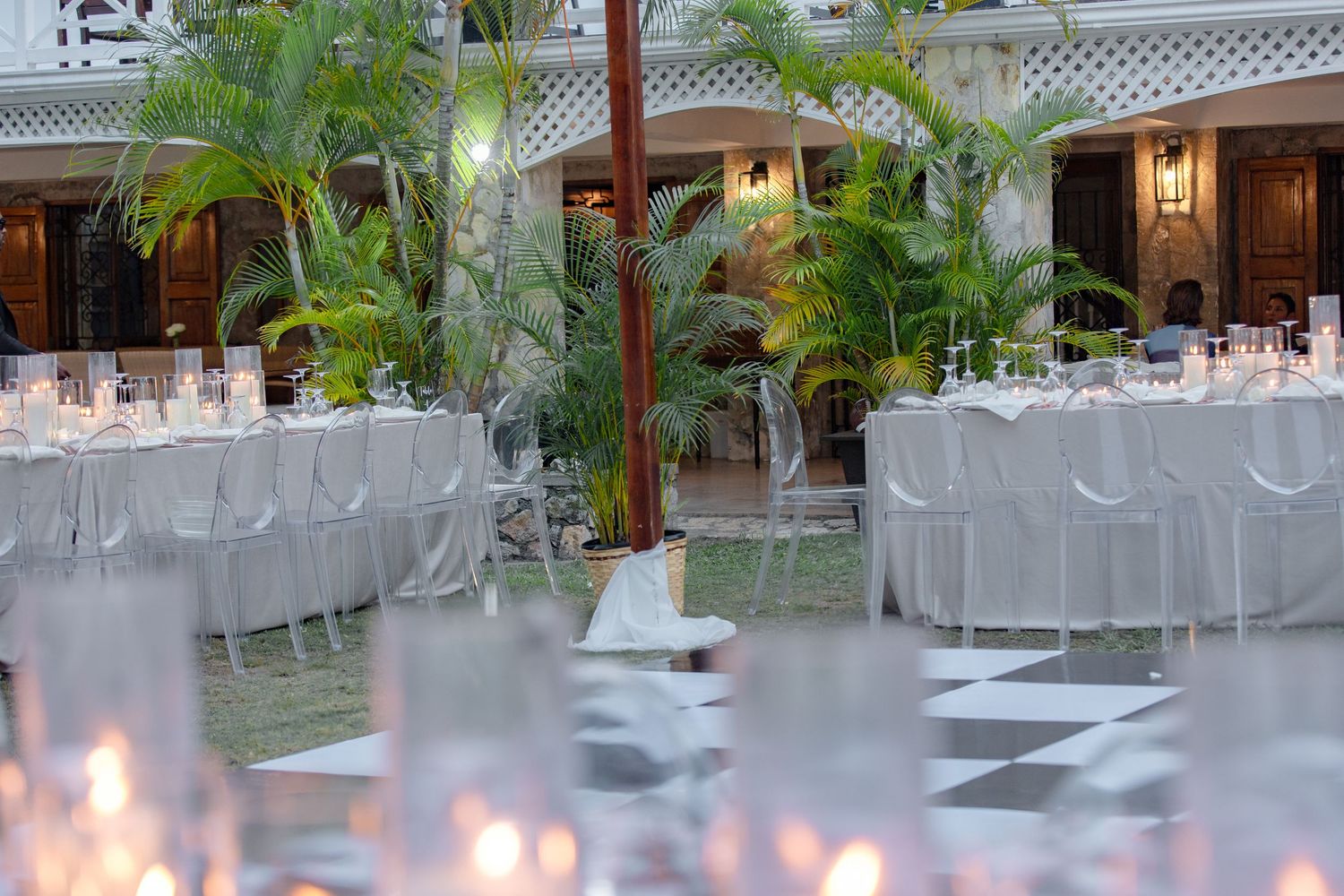 Elegant outdoor wedding reception with white draped tables, ghost chairs, tropical plants, and glowing candles.