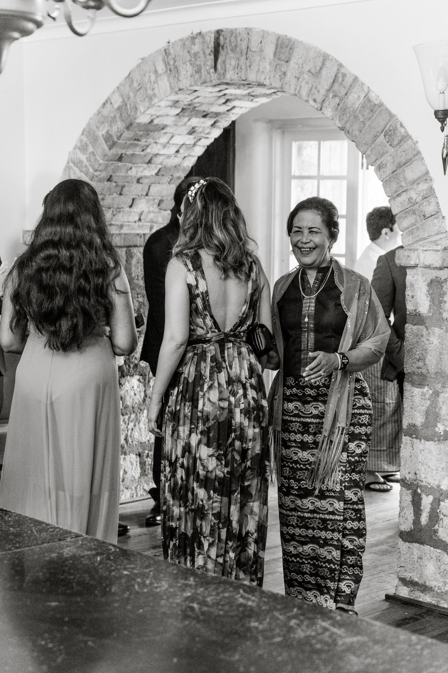 Black and white photo of women in traditional Myanmar attire conversing under a rustic brick archway at a wedding.