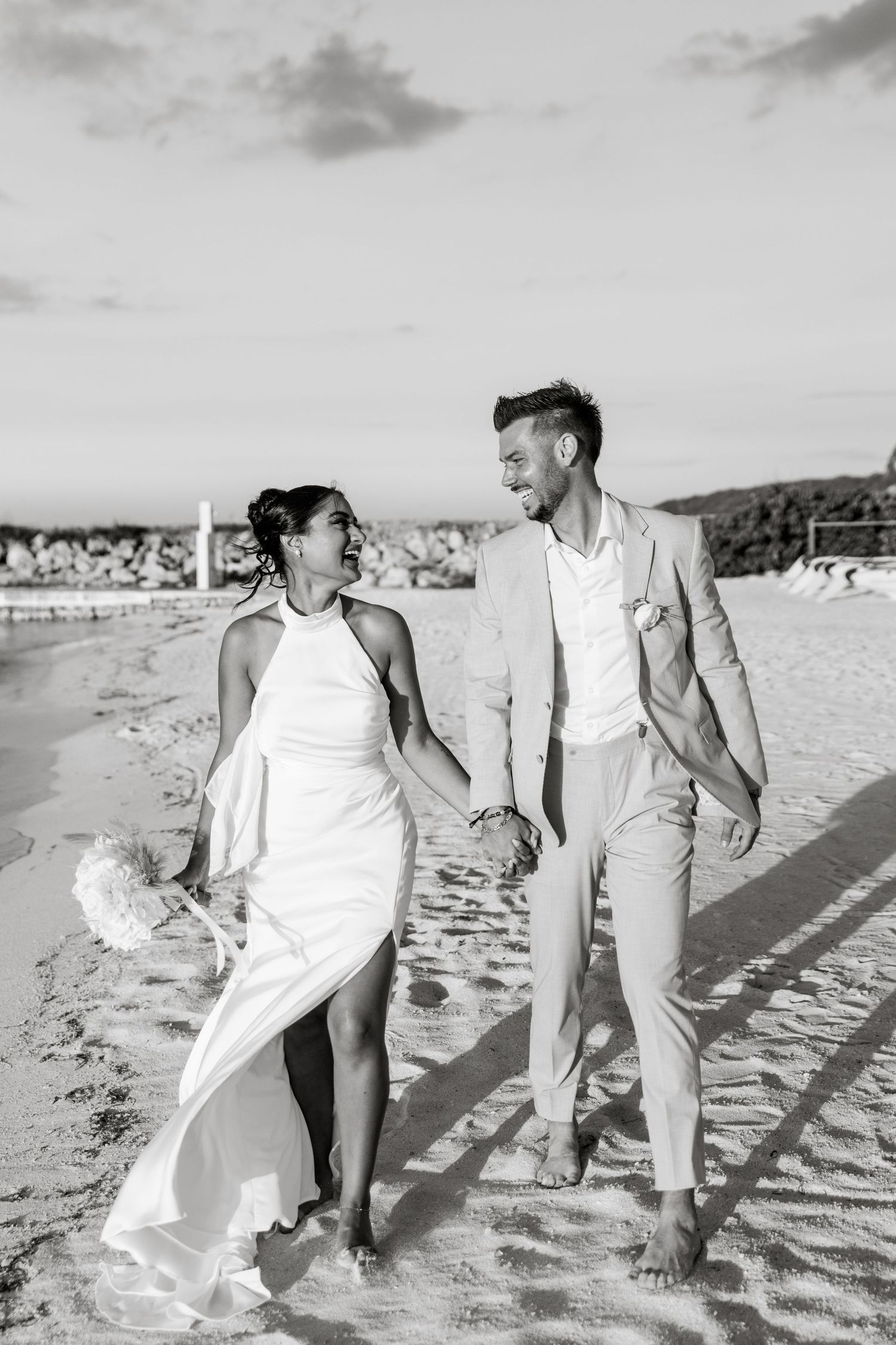 Black and white photo of newlyweds holding hands and walking barefoot on a sandy beach at sunset.
