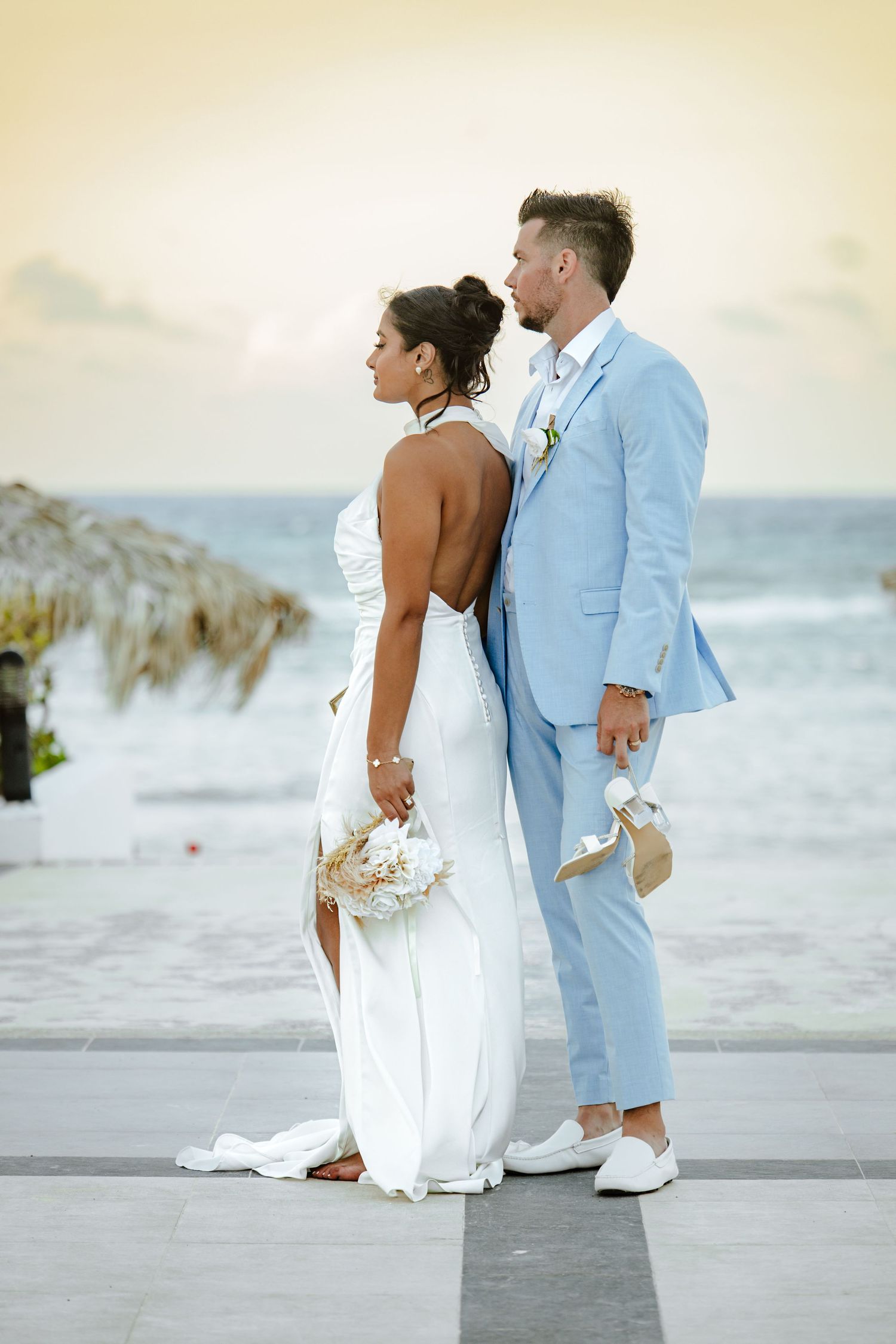 Bride in white backless gown and groom in light blue suit pose together by the ocean at a beach wedding.