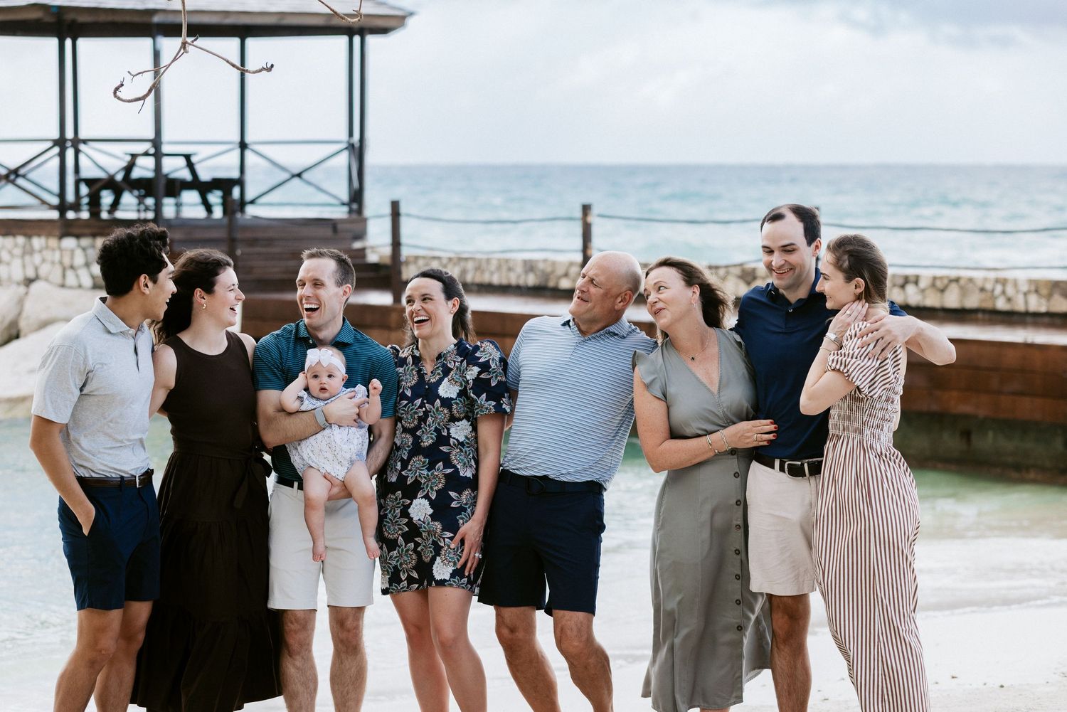 Happy family group of eight adults and a baby laughing together on a tropical beach near a wooden pier.