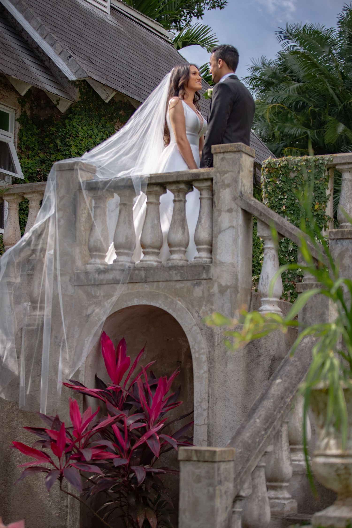 Wedding couple stands on ornate stone balcony surrounded by tropical greenery and red plants.