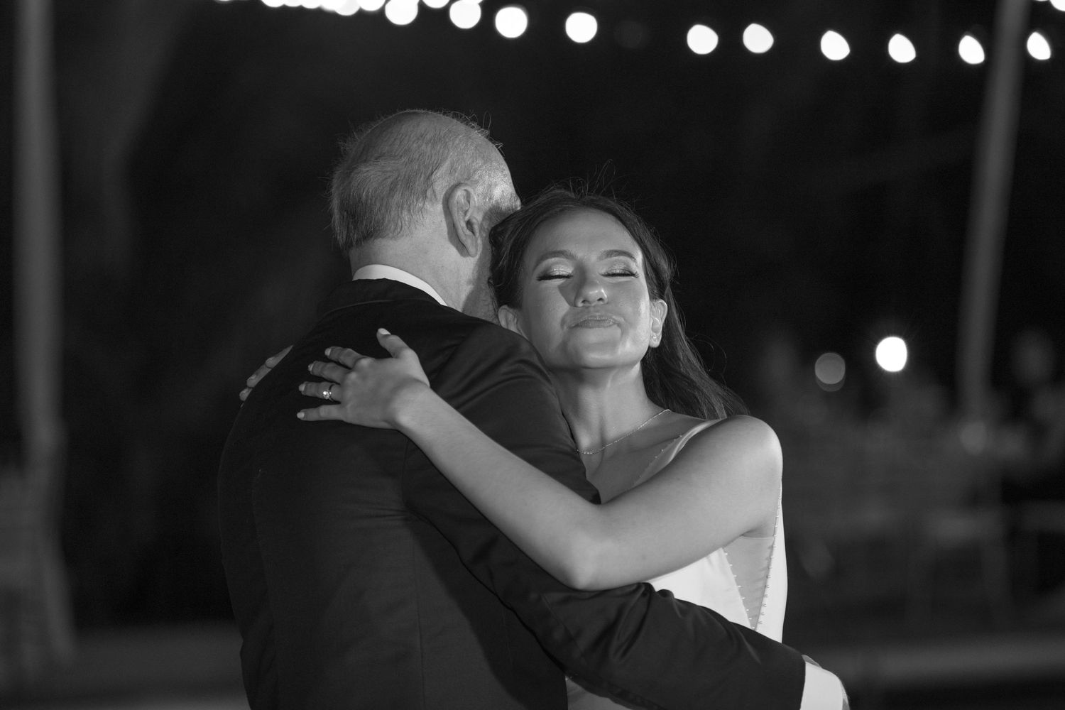 Black and white photo of bride and groom sharing an emotional first dance under string lights at night.