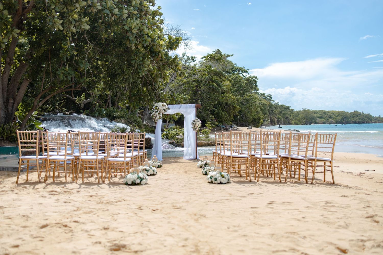 Elegant beach wedding ceremony setup with gold chiavari chairs and white floral arch on sandy shore.