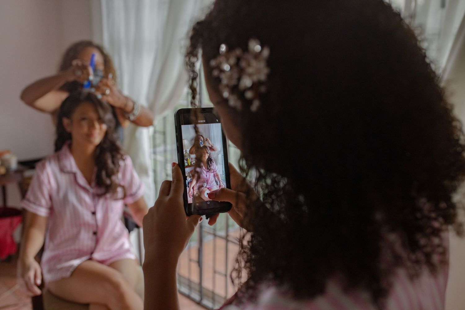 Bridesmaid taking a photo on smartphone of bride getting hair done in bright bedroom.