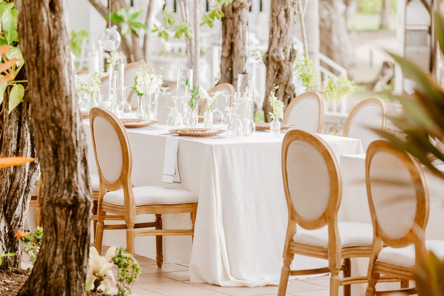 Elegant outdoor wedding reception table with white linen, gold chairs, candles, and greenery decor.