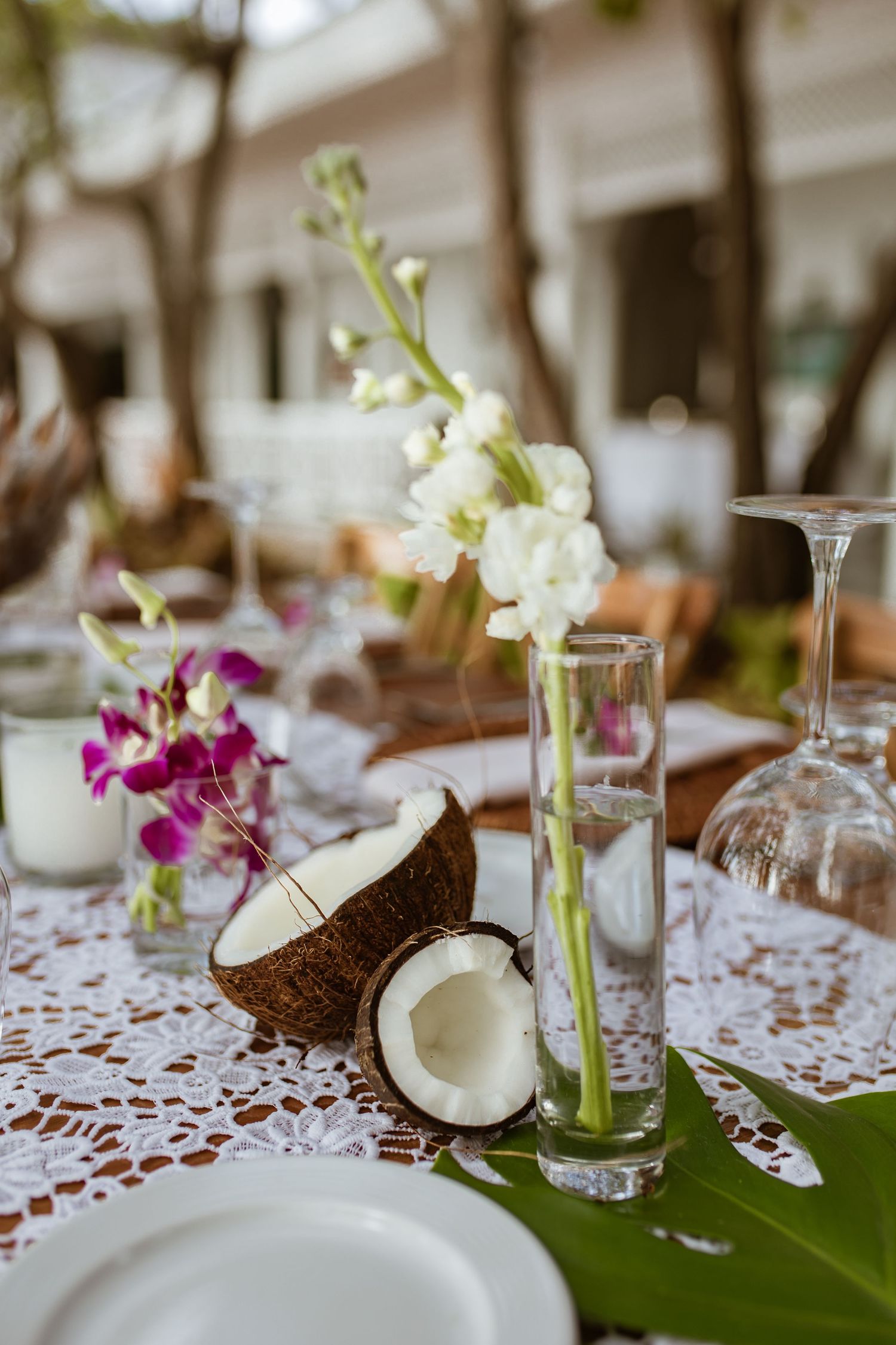 Tropical wedding table setting with split coconut, white orchids, purple flowers, and lace tablecloth outdoors.