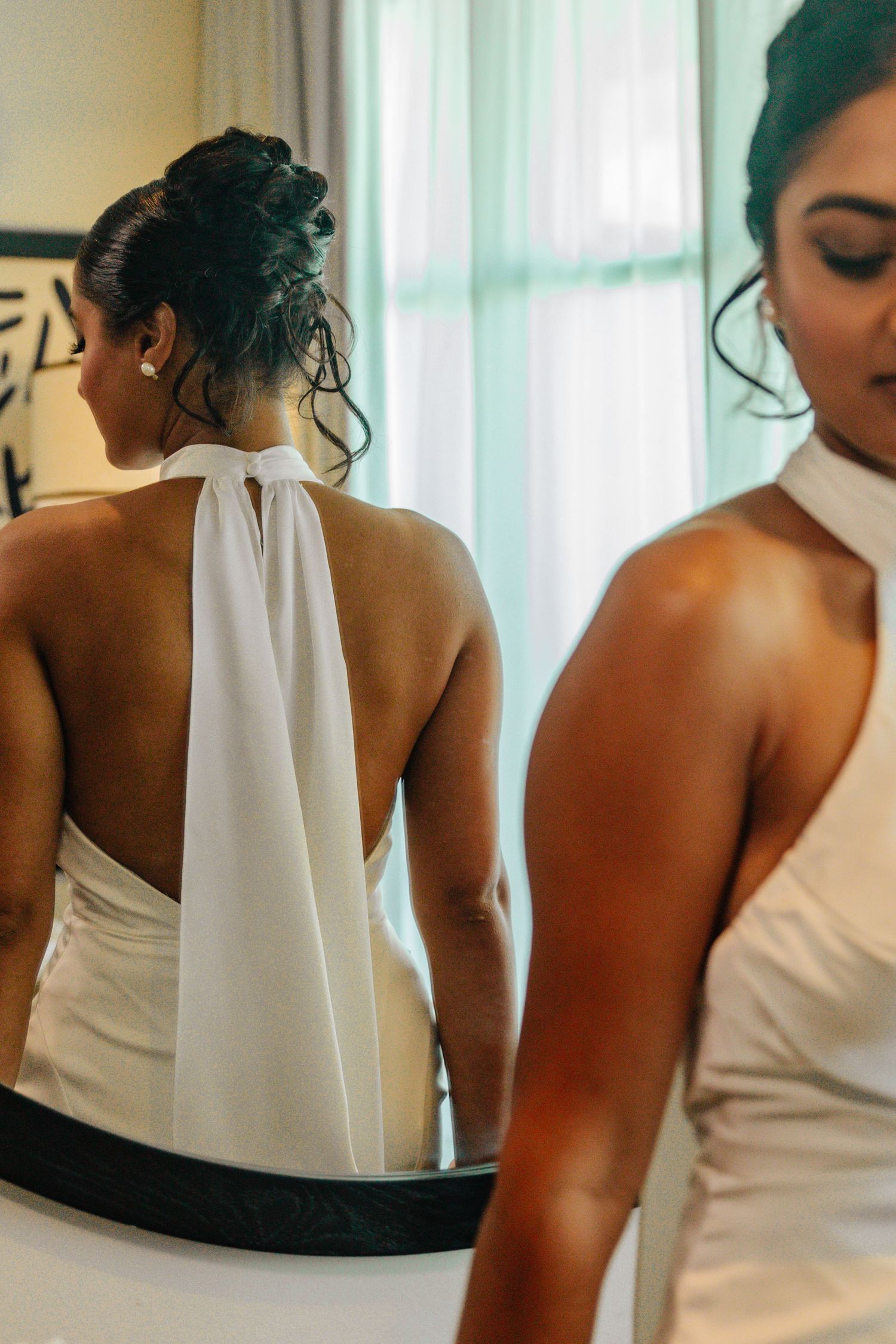 Bride in white halter-neck wedding dress with open back, viewed from behind in mirror reflection.