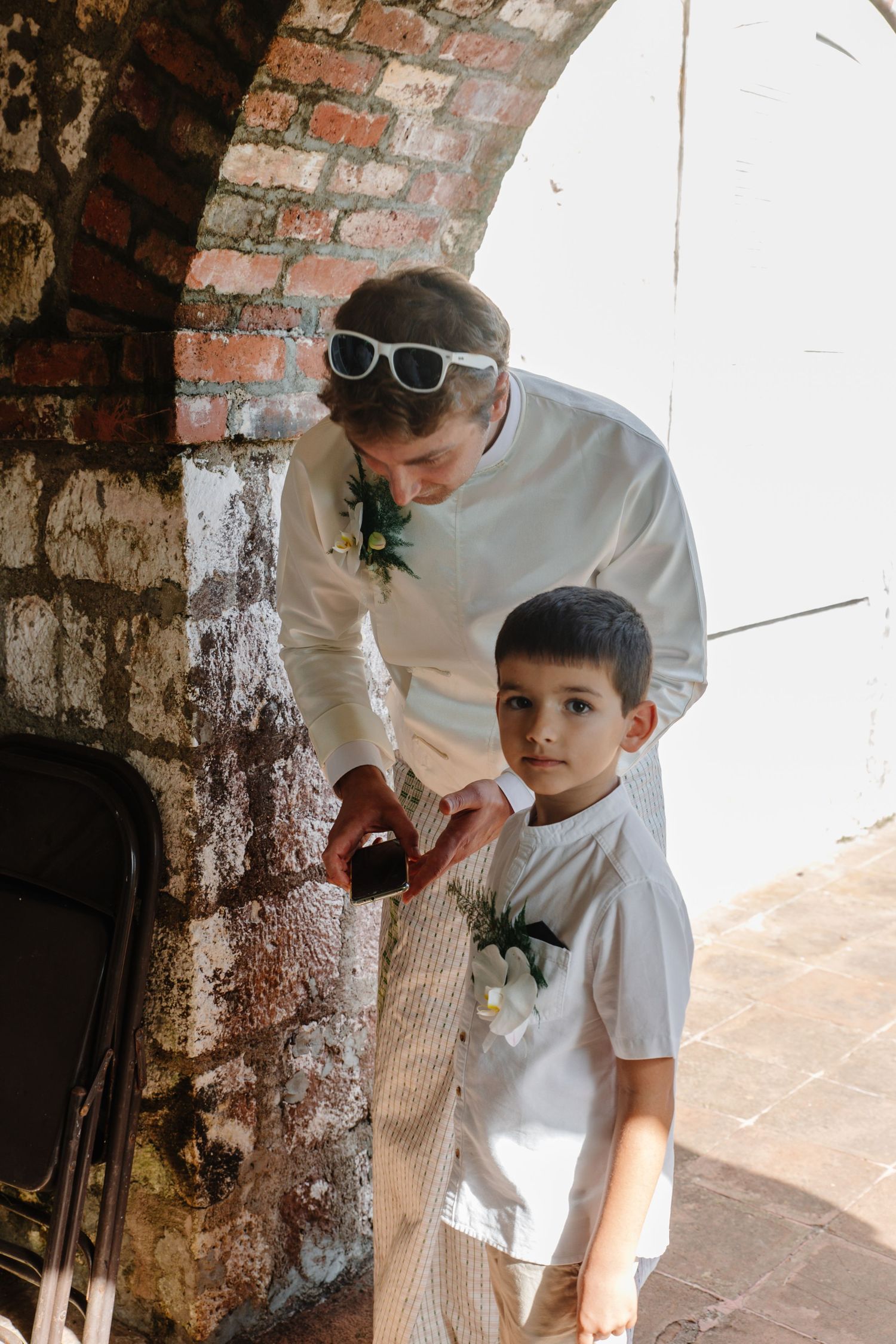 Man in white wedding attire with sunglasses assists young boy with boutonniere under rustic brick archway.