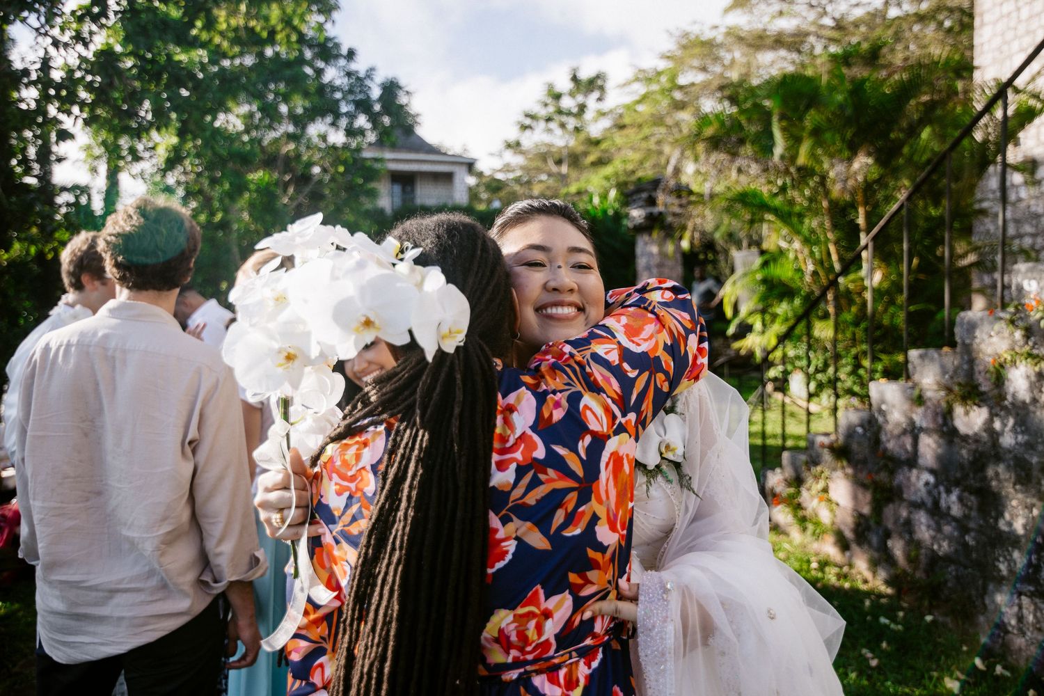 Two women hugging and smiling at outdoor wedding, one holding white orchid bouquet.