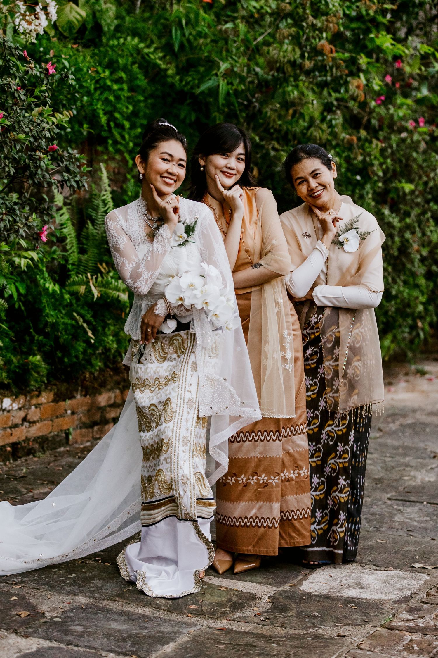 Bride in traditional white embroidered gown poses with two smiling bridesmaids in a lush garden.
