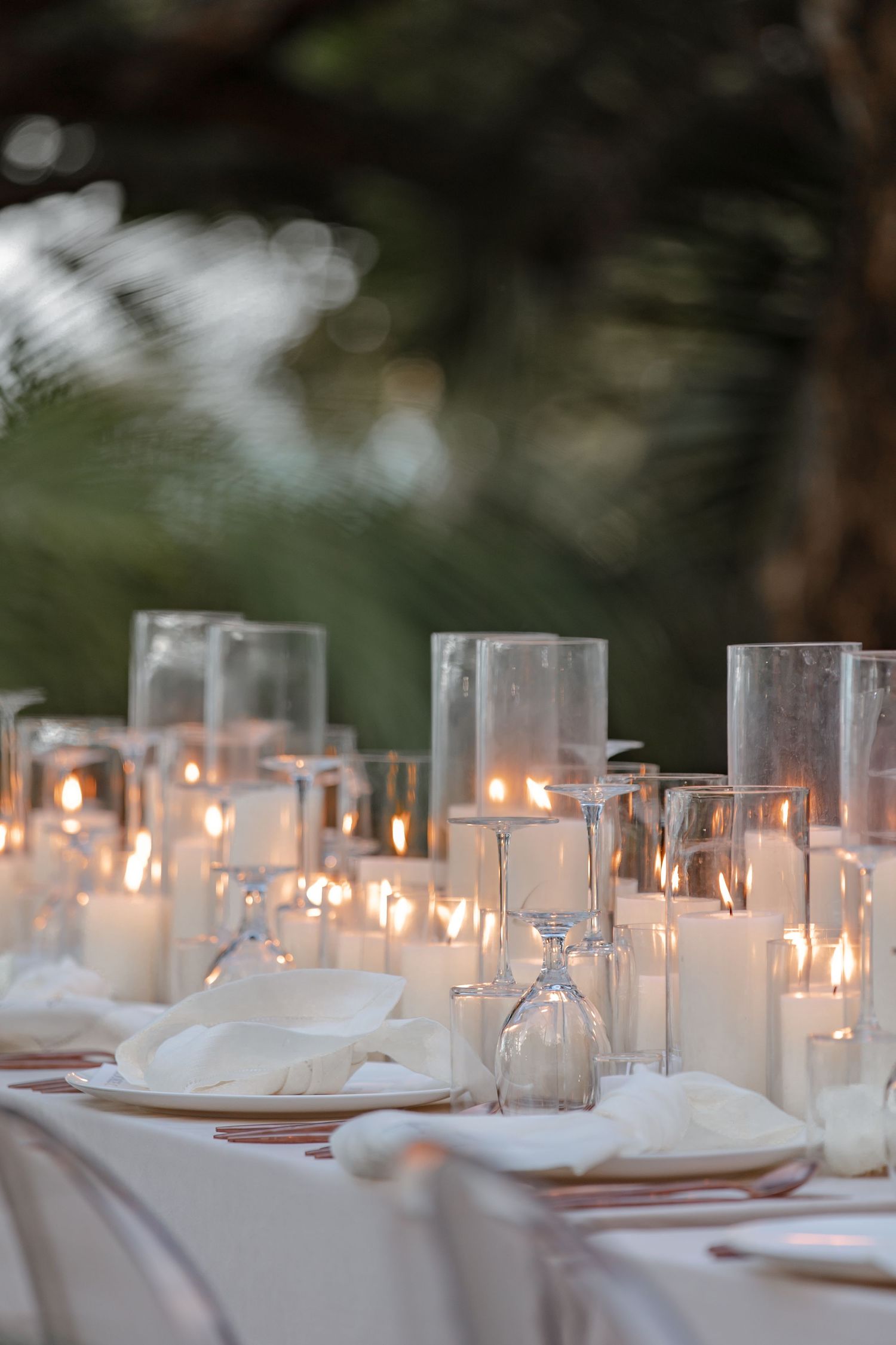 Elegant wedding reception table with glowing candles in glass holders and white place settings at dusk.