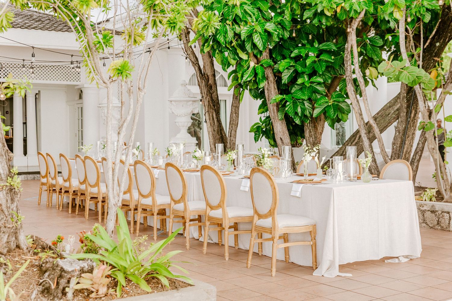 Outdoor garden wedding reception with long white linen tables, gold chiavari chairs, and lush green tree backdrop.