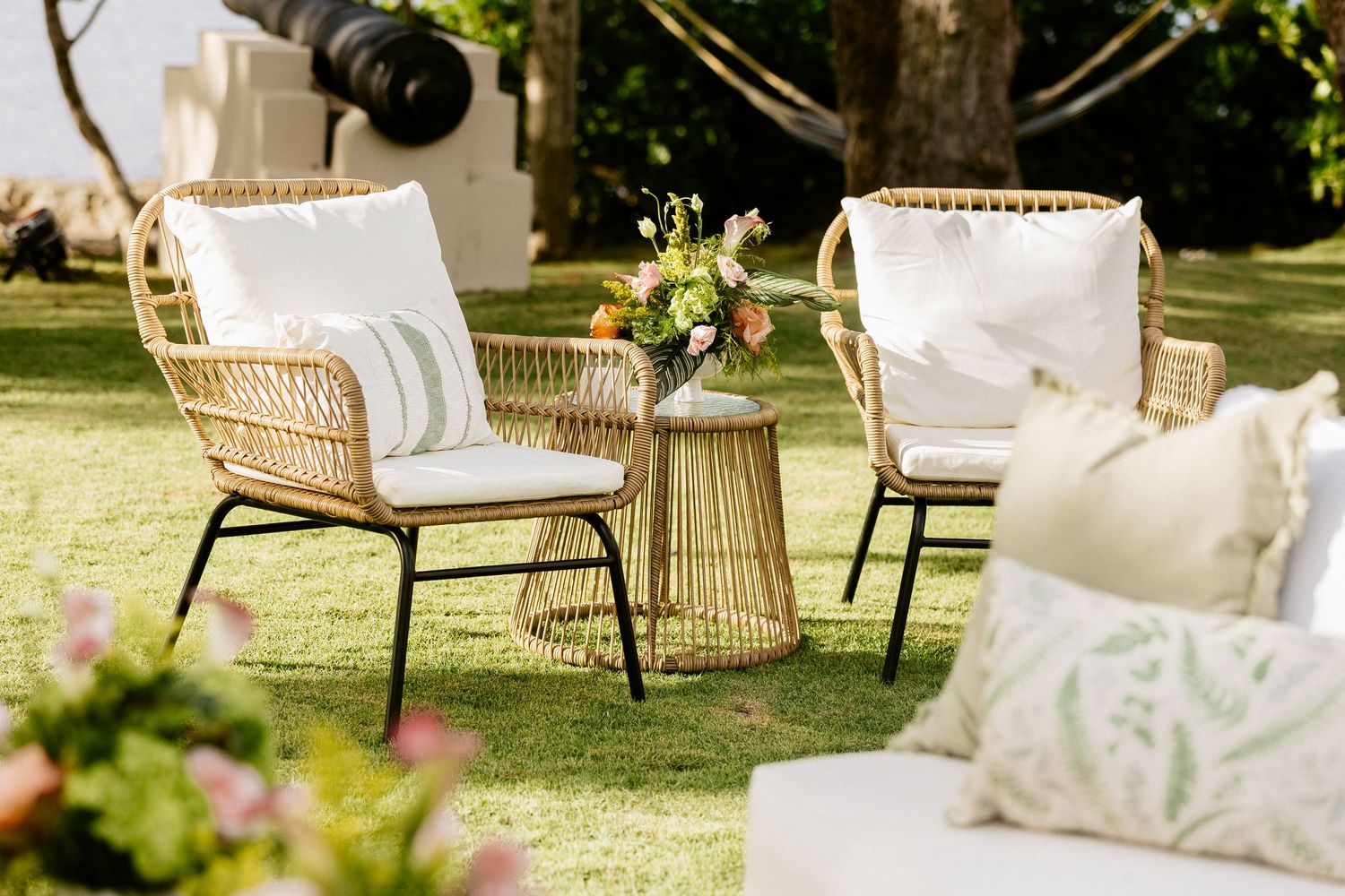 Two wicker chairs with white cushions and a small round table with flowers on a green lawn.