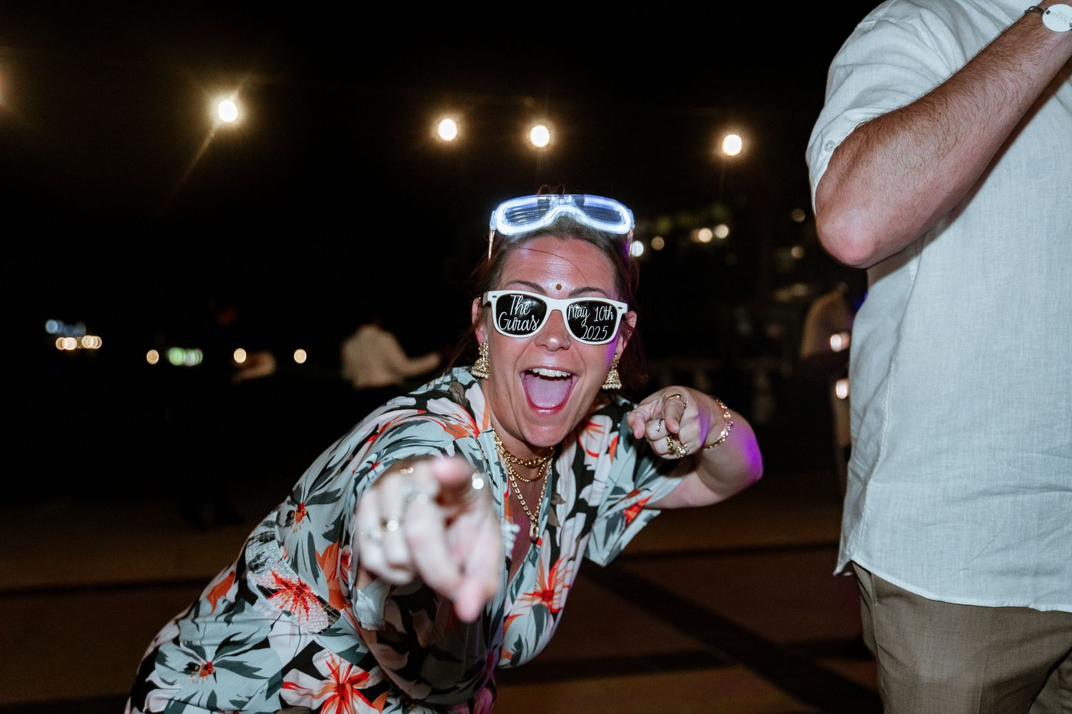 Excited woman in floral shirt and sunglasses dancing at outdoor nighttime wedding reception.