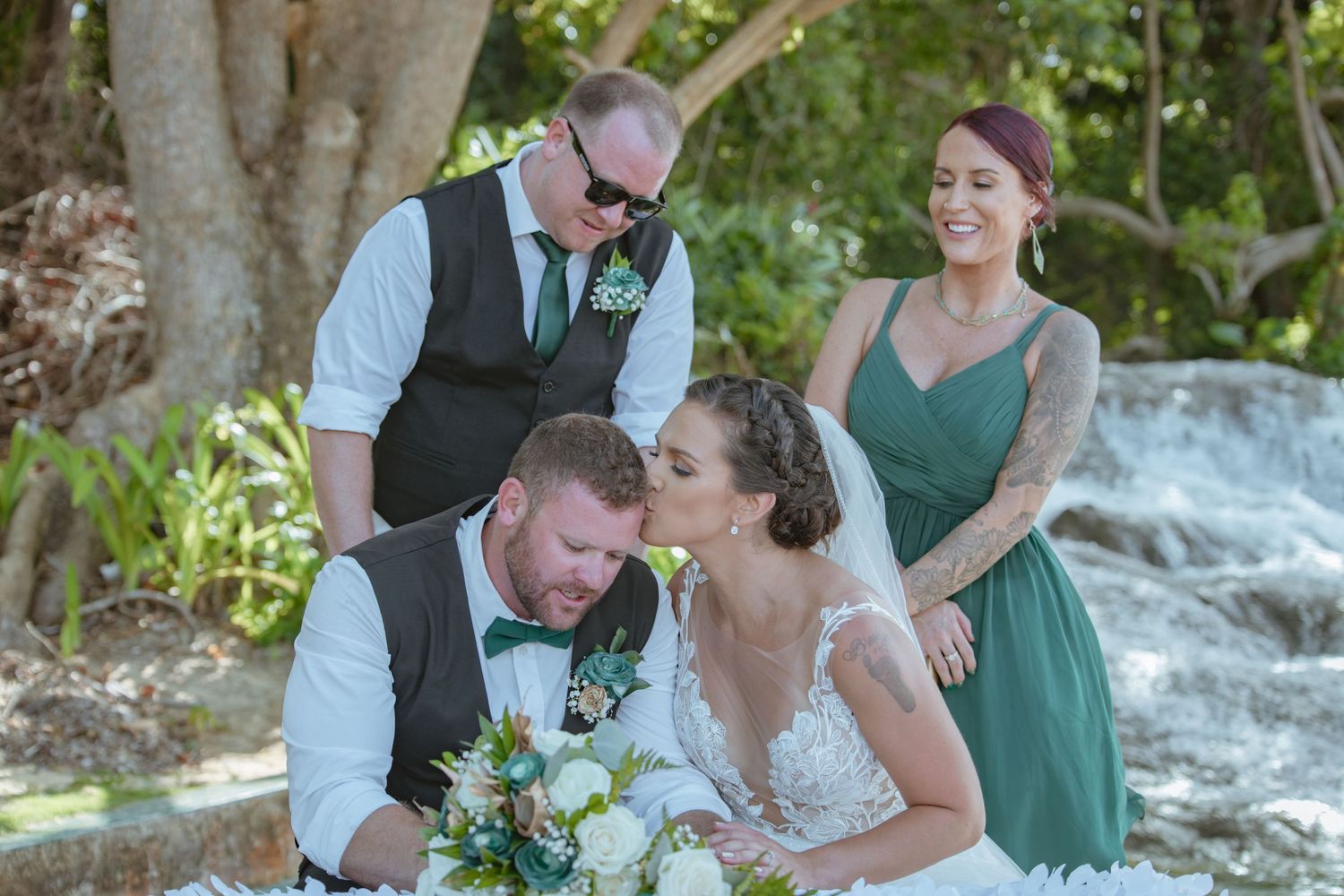 Bride and groom laugh with wedding party beside a waterfall in tropical garden setting.