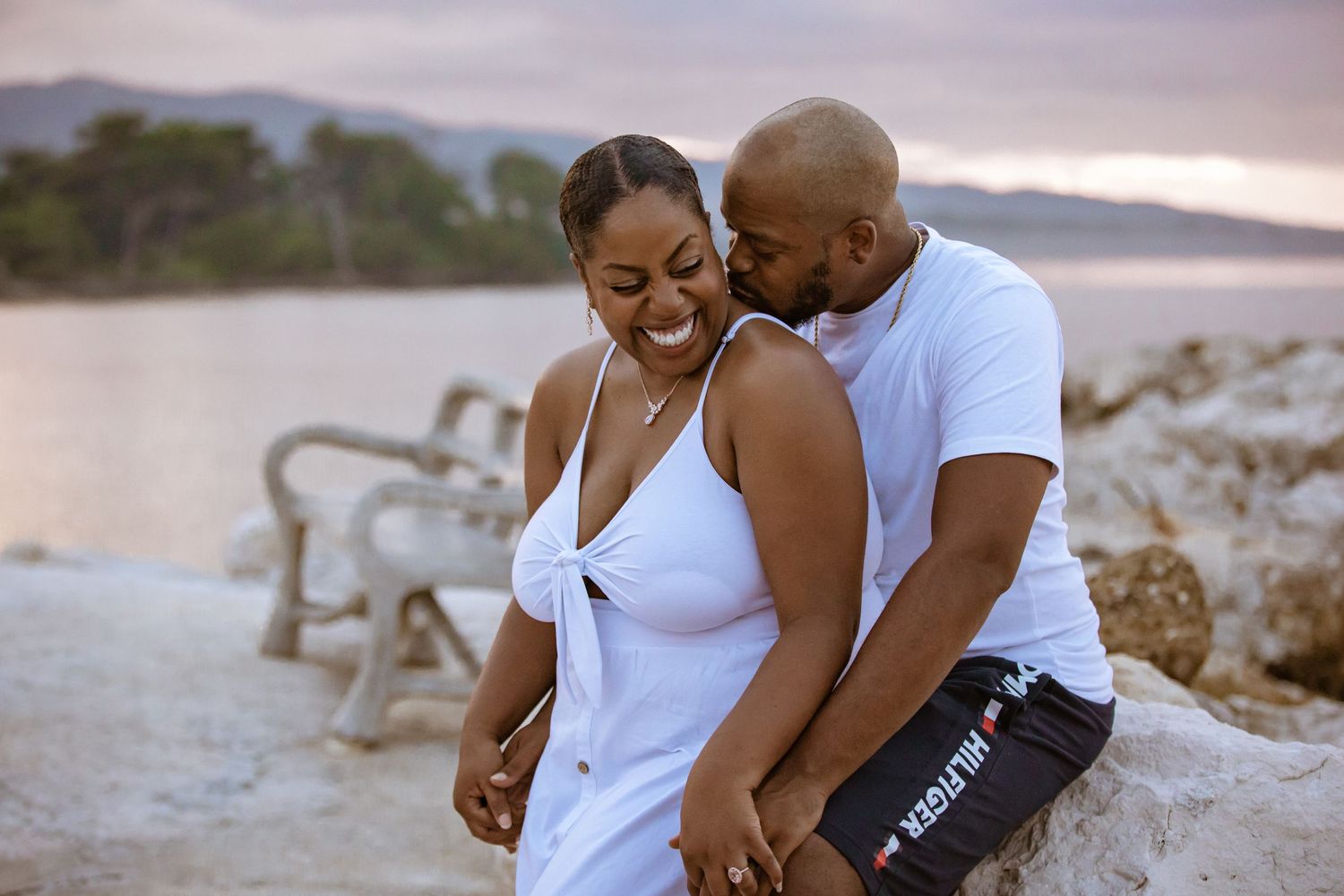 Happy couple sharing a romantic moment by the waterfront at sunset, dressed in white casual attire.