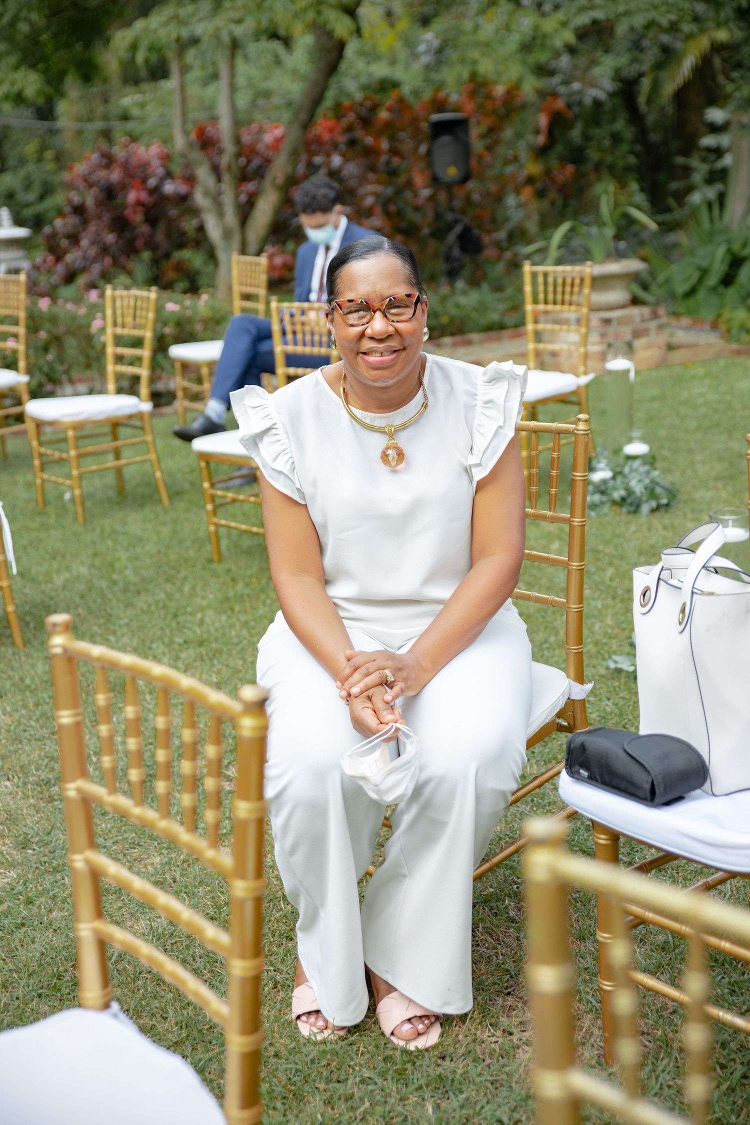 Smiling woman in white outfit seated on gold chiavari chair at outdoor wedding ceremony.