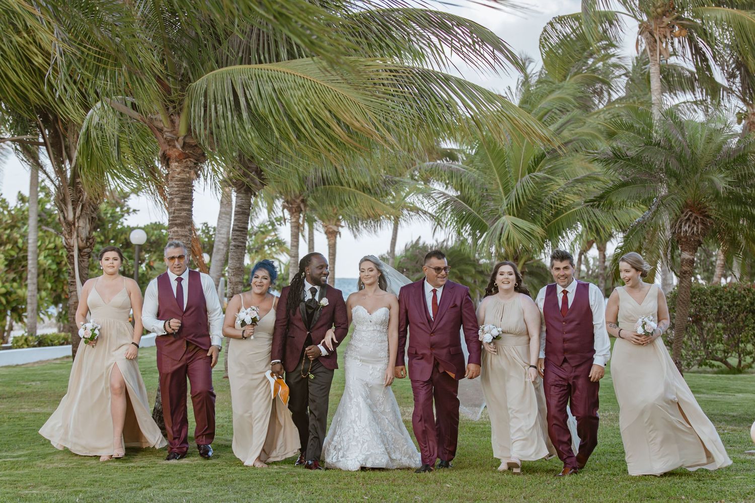 Wedding party walks together on tropical lawn with palm trees, wearing burgundy and champagne attire.