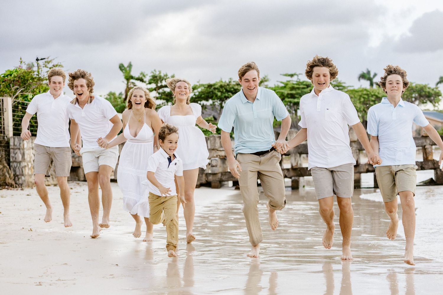 Large family group running joyfully on a tropical beach, wearing coordinated white and khaki outfits on a cloudy day.