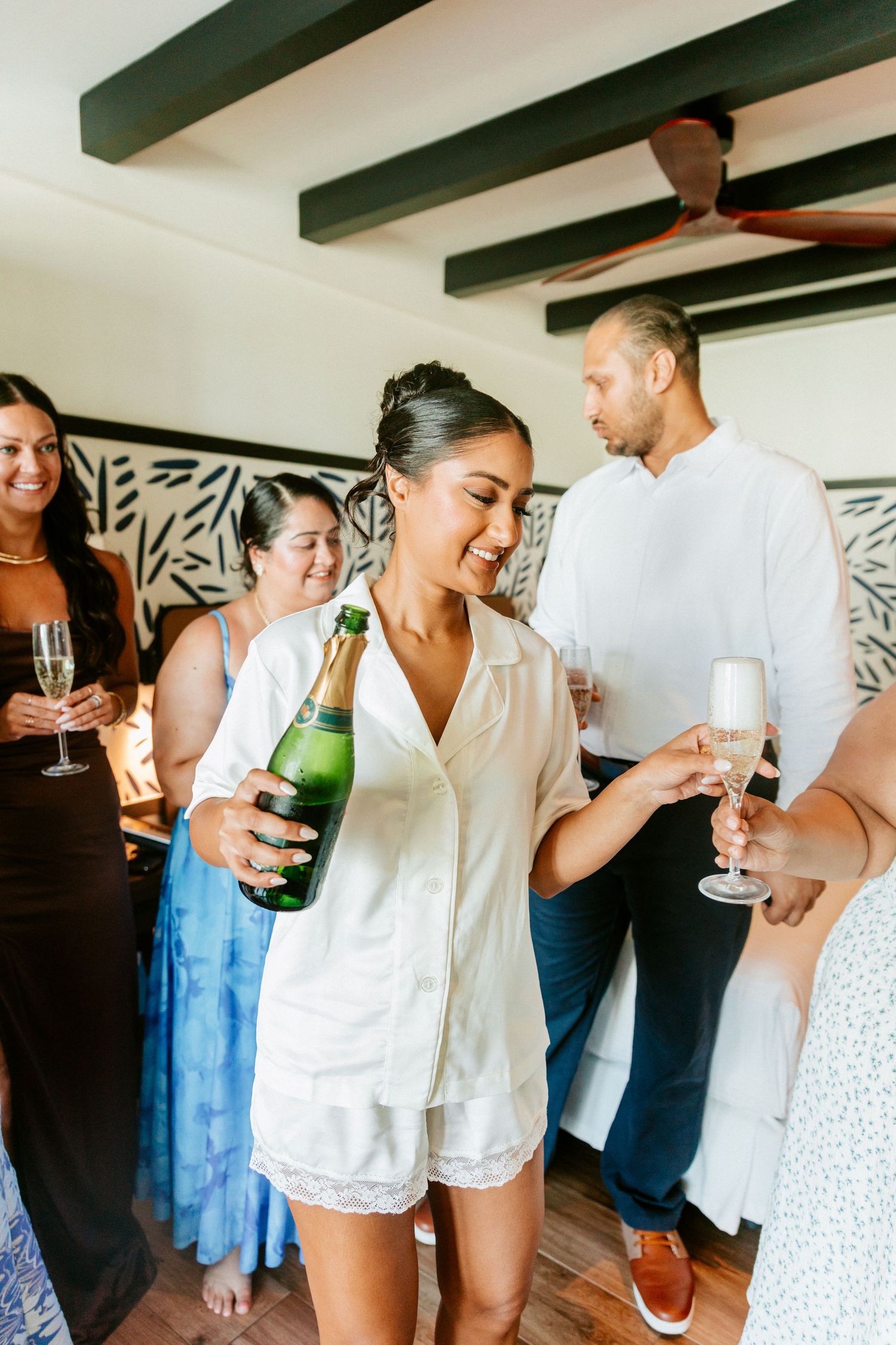 Bride in white pajamas pours champagne for friends during wedding morning preparations in a tropical resort room.