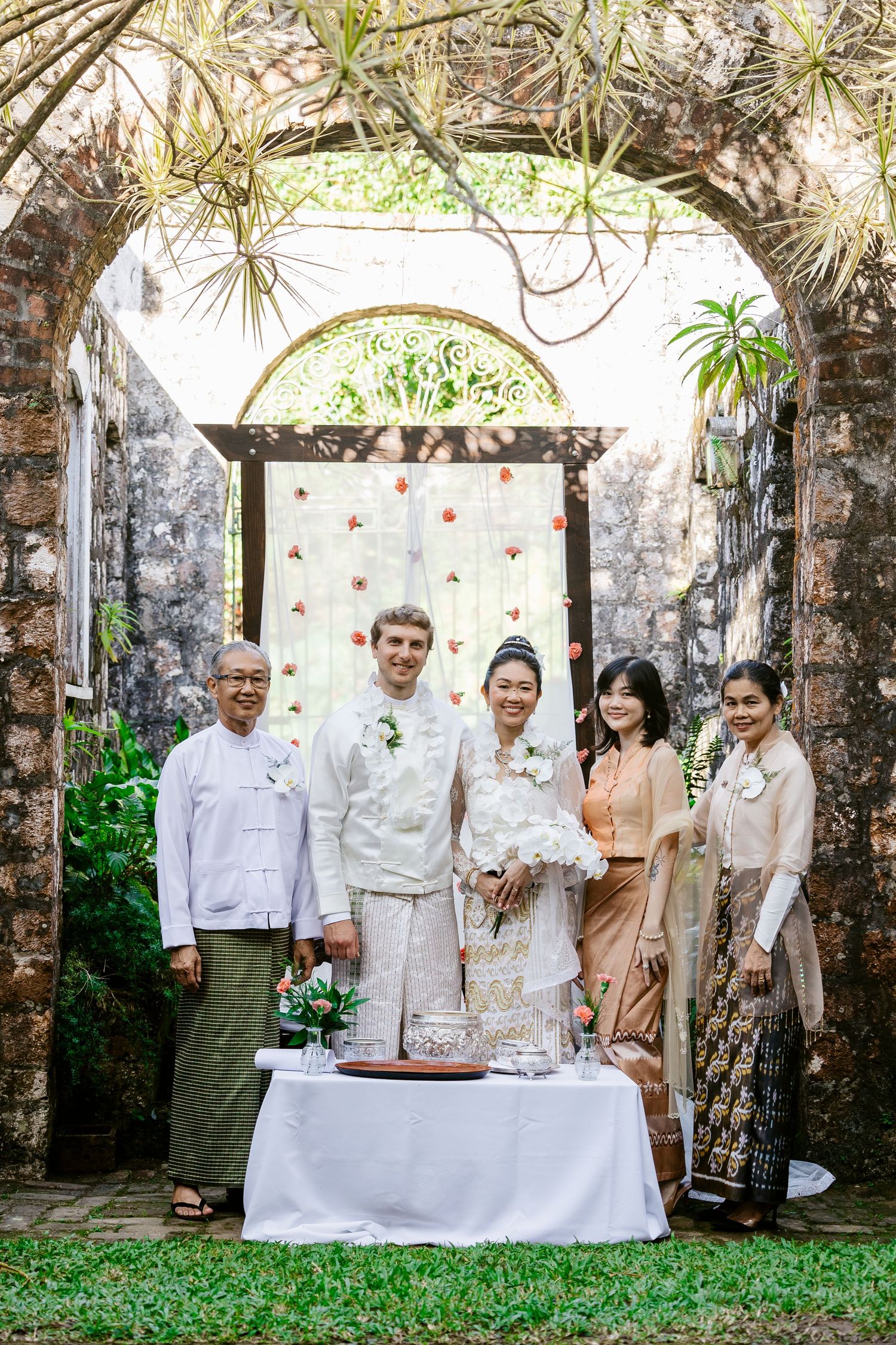 Couple in traditional Myanmar wedding attire pose with family under stone arch with floral backdrop outdoors.